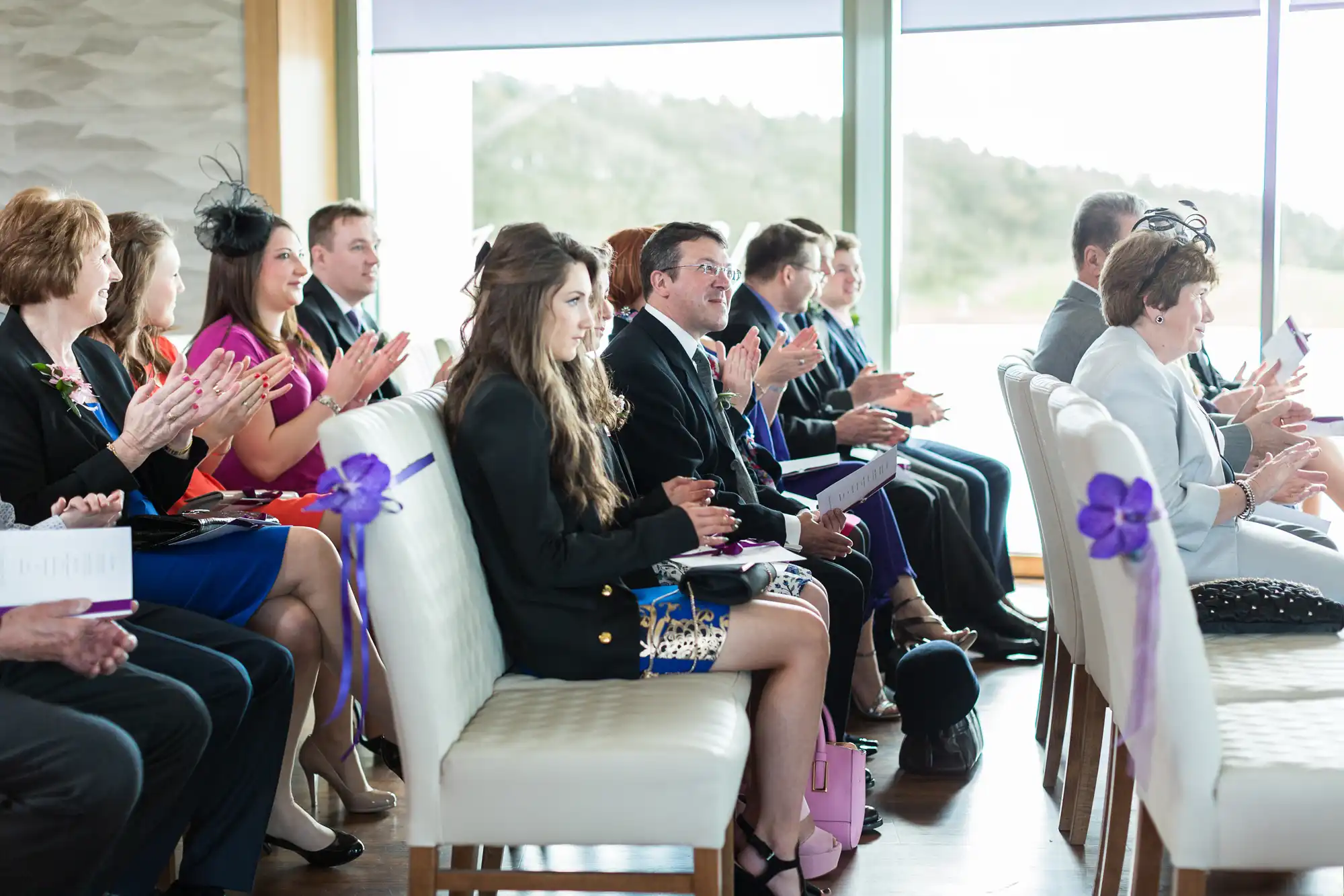Guests at a formal event, seated in rows, clapping and holding programs, with a venue featuring large windows and a view.