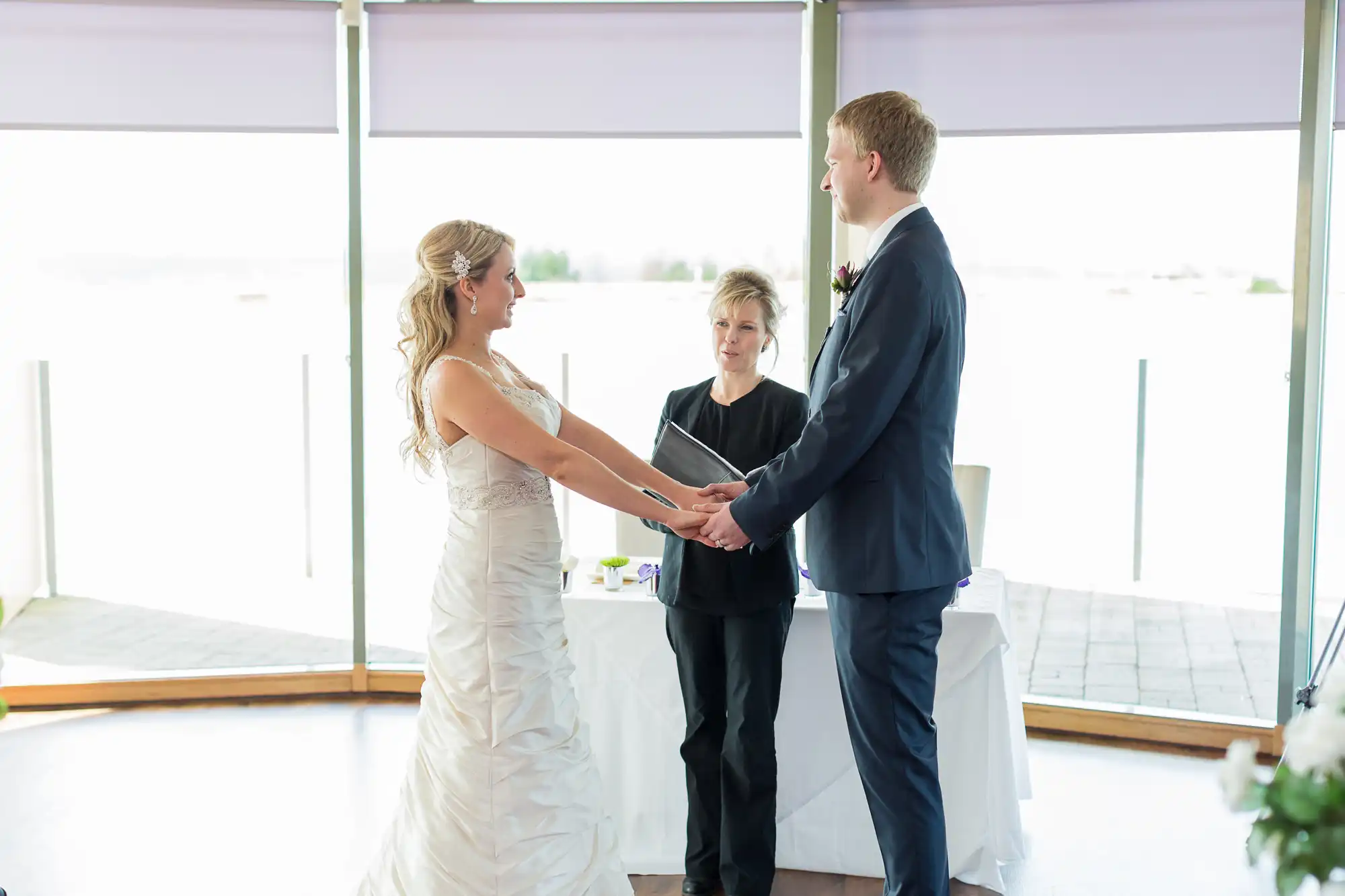 Bride and groom holding hands during wedding ceremony, with an officiant standing between them, inside a bright venue with large windows.