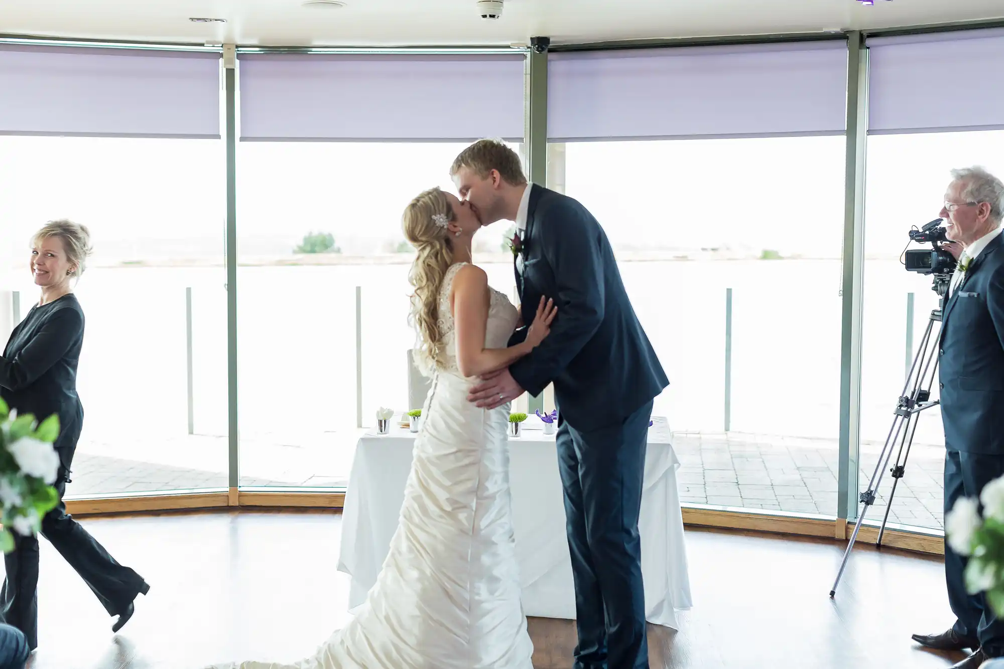 A bride and groom kissing in a room with large windows, while a photographer captures the moment and a woman watches smiling.