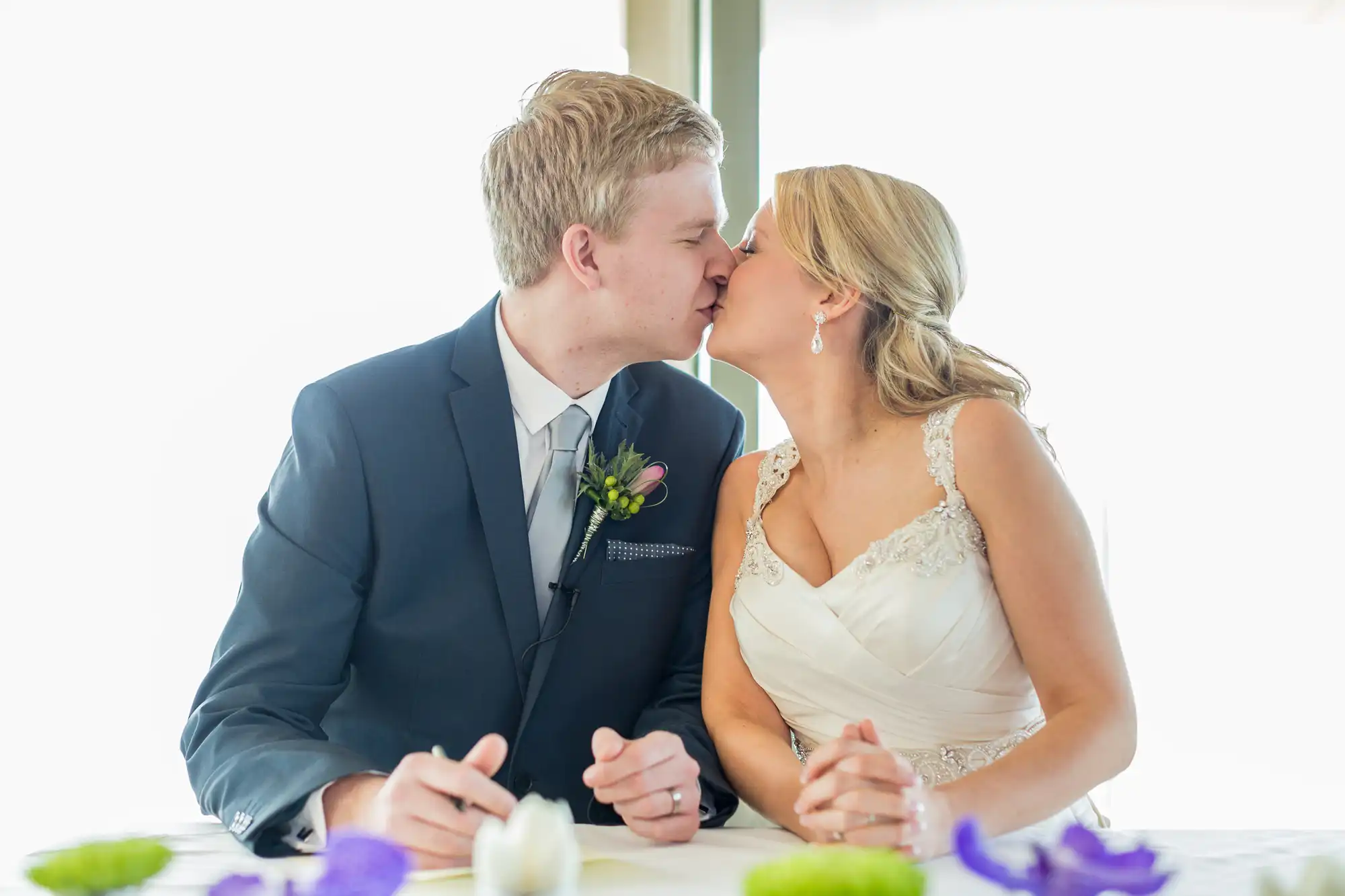 A bride and groom kissing at their wedding reception table, dressed in formal attire, with a bright window backlighting them.