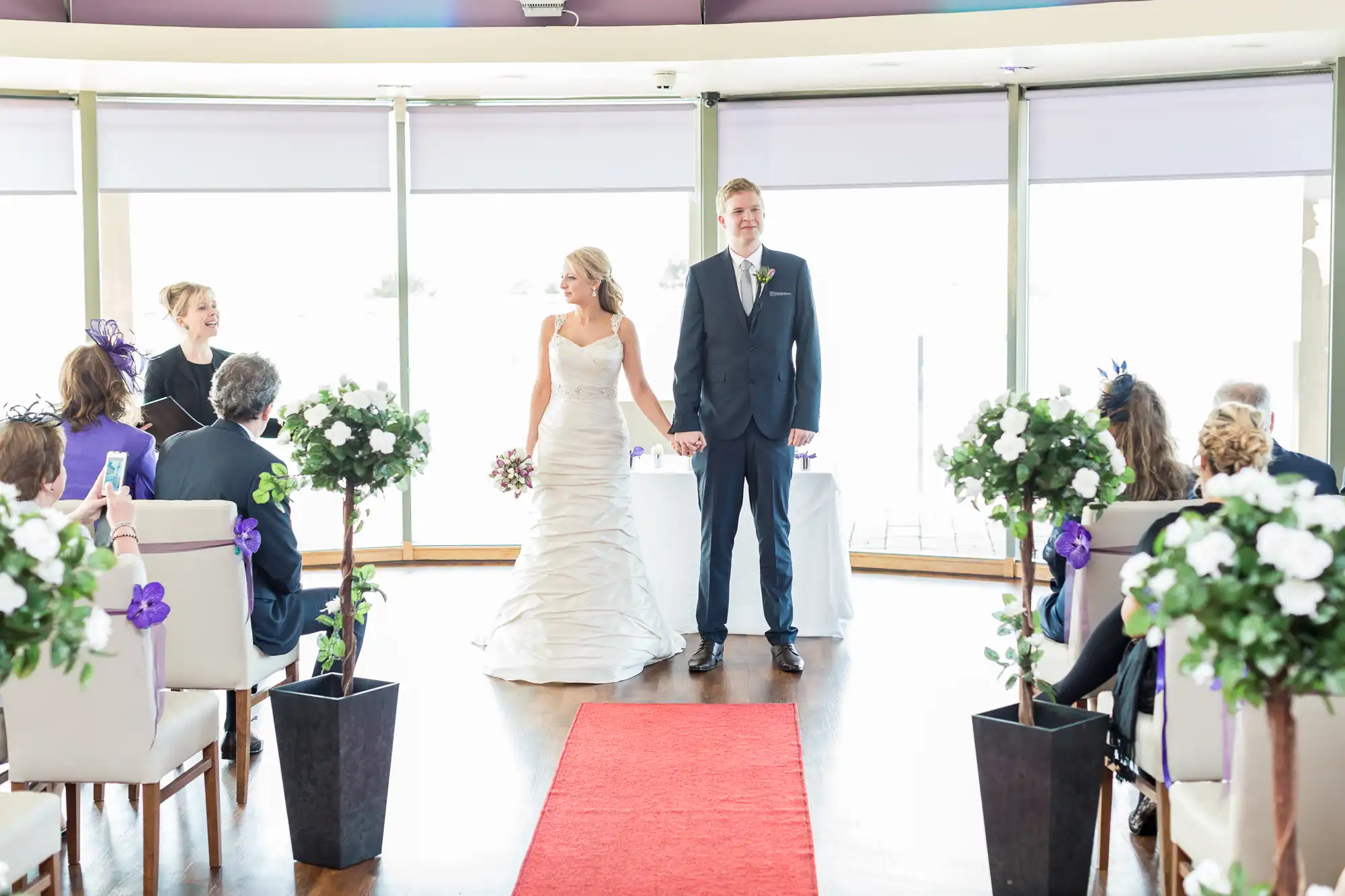 A bride and groom hold hands while walking down the aisle during their wedding ceremony, surrounded by seated guests in a venue with large windows.