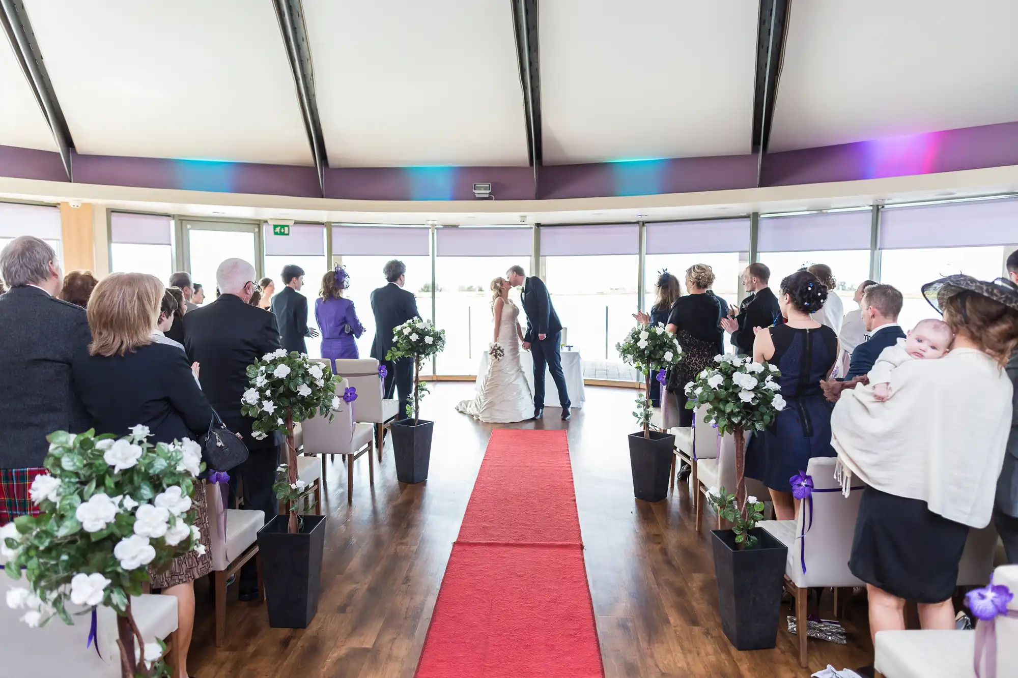 Wedding ceremony in a tent with guests facing a couple exchanging vows at the end of a red carpet, surrounded by floral decorations.