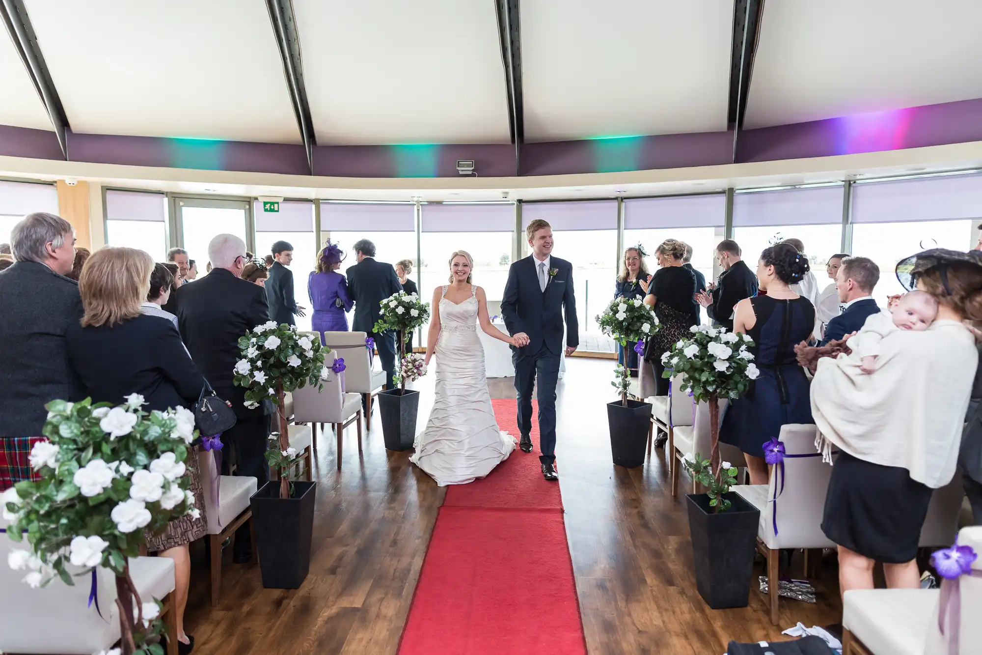 A bride and groom walk down the aisle smiling, surrounded by guests in a modern venue with large windows and purple lighting.