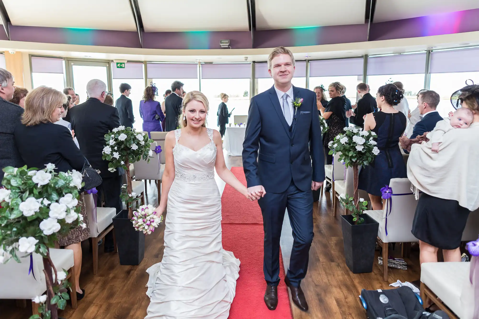 A bride and groom holding hands, smiling as they walk down the aisle in a room with guests standing around them.