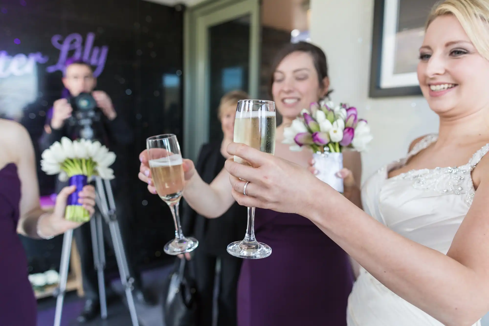 A bride in a white dress toasts with champagne, flanked by bridesmaids in purple, with a photographer capturing the moment.