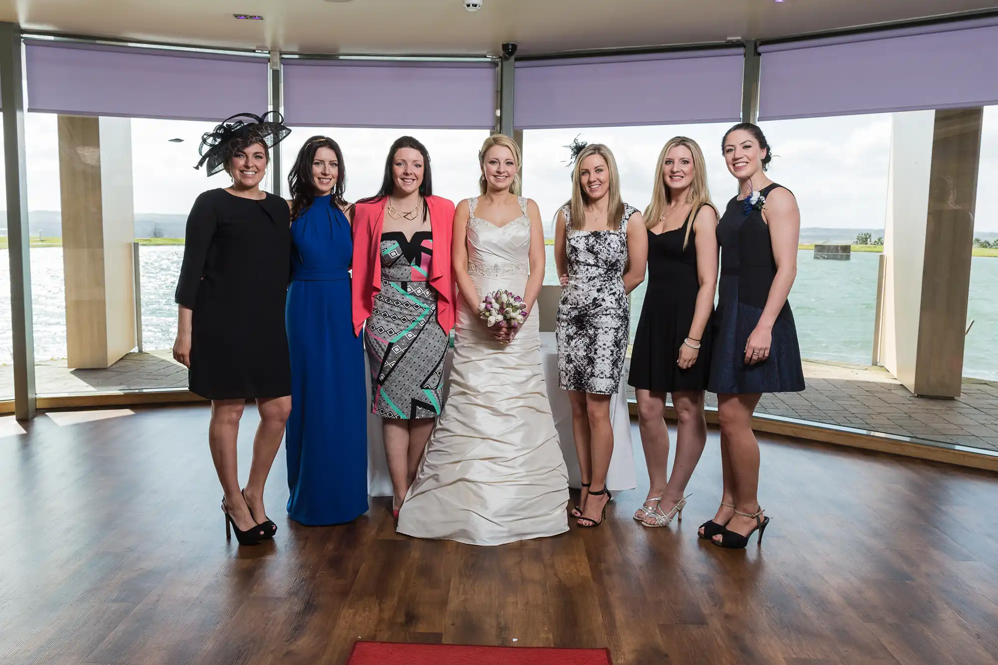 Seven women, including a bride, pose together indoors in front of large windows overlooking a body of water. they are dressed in a mix of colorful formal attire and smiles.