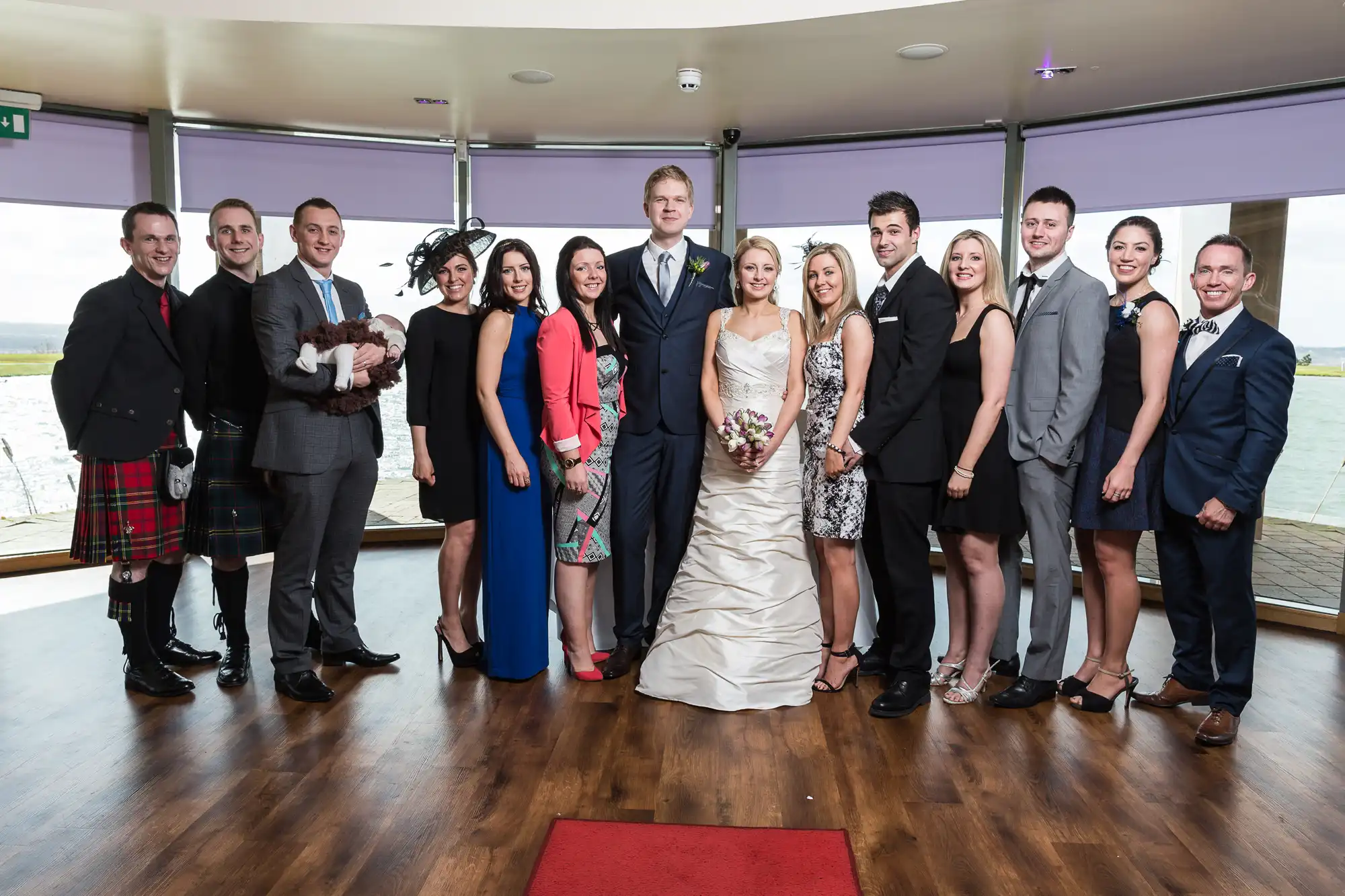 A group of elegantly dressed guests posing with a bride and groom in a room with large windows overlooking a body of water.