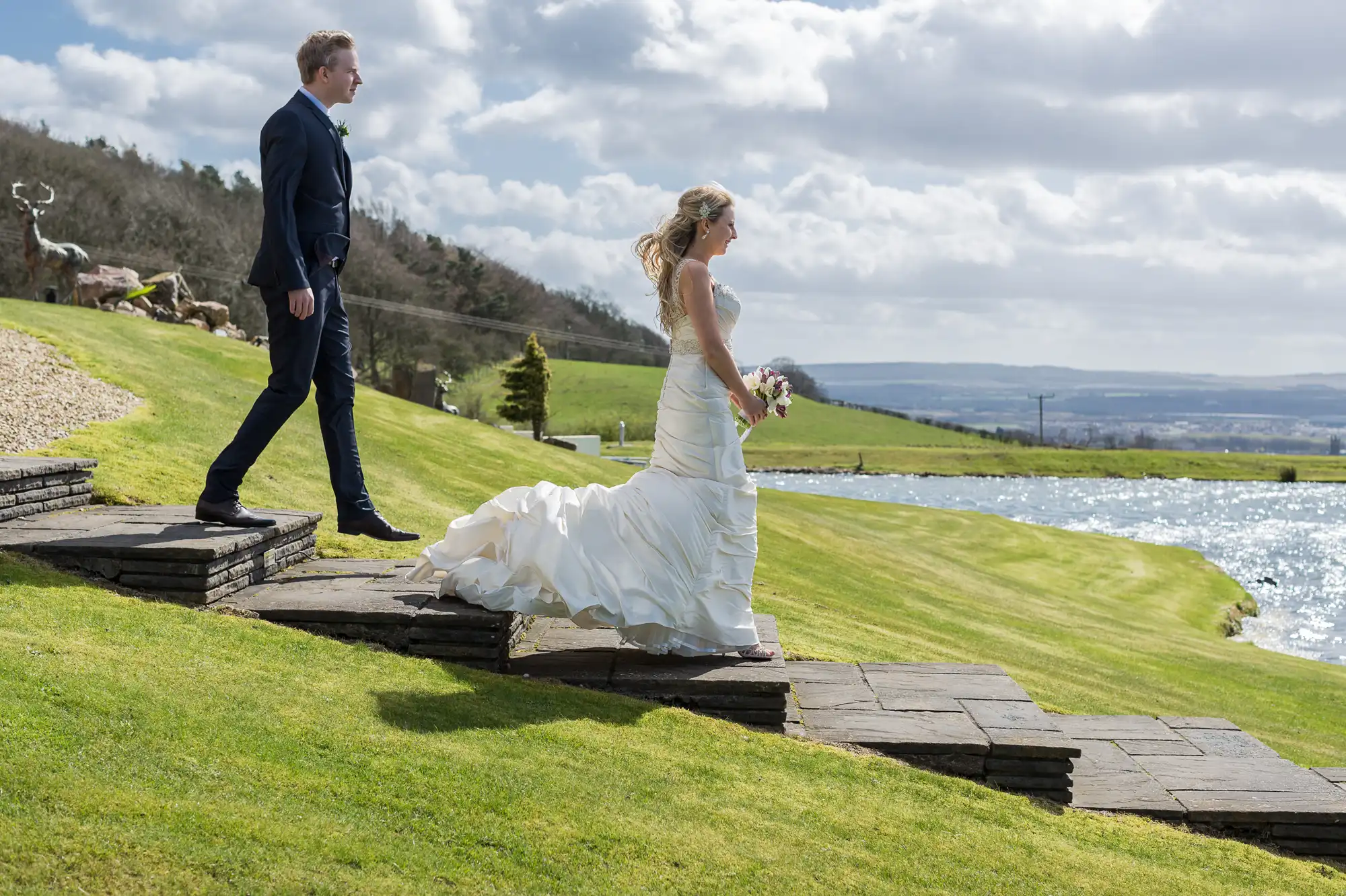 Bride and groom walking on a stone path beside a lake with rolling hills in the background under a partly cloudy sky.