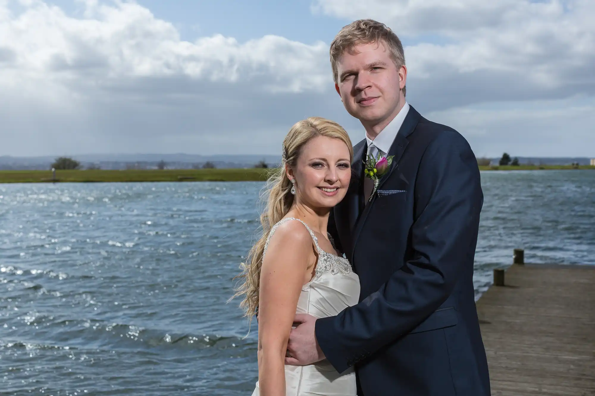 A bride and groom embracing on a wooden pier, with a serene lake and cloudy sky in the background.