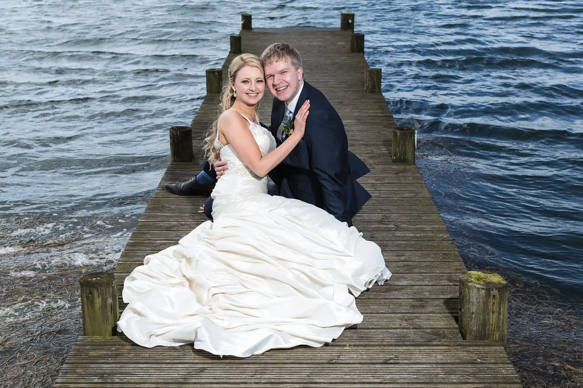 Bride and groom sitting on a wooden jetty by a lake, smiling at the camera, bride in a flowing white gown and groom in a dark suit.