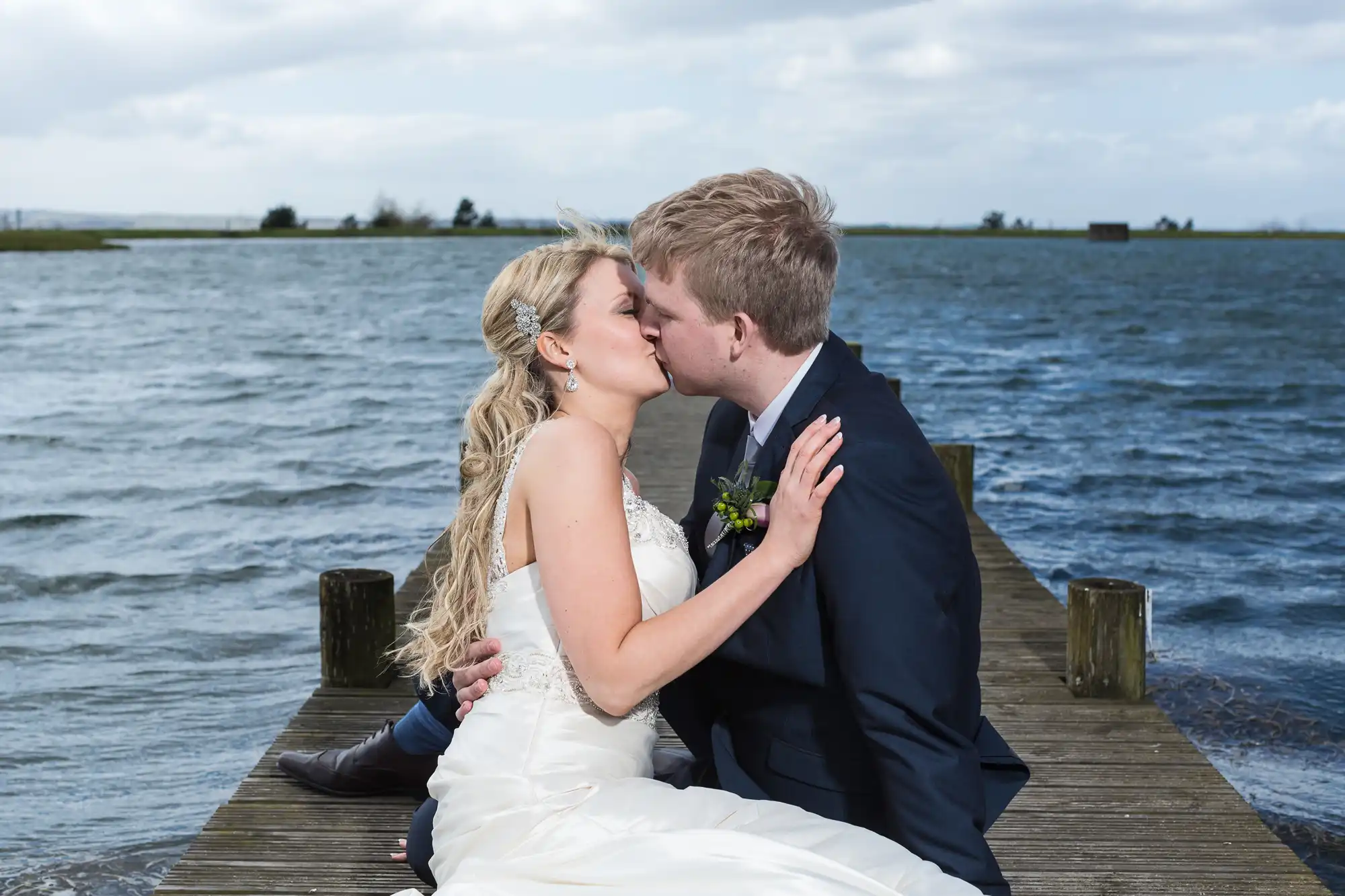 A bride and groom kissing on a wooden dock by a lake, under a cloudy sky.