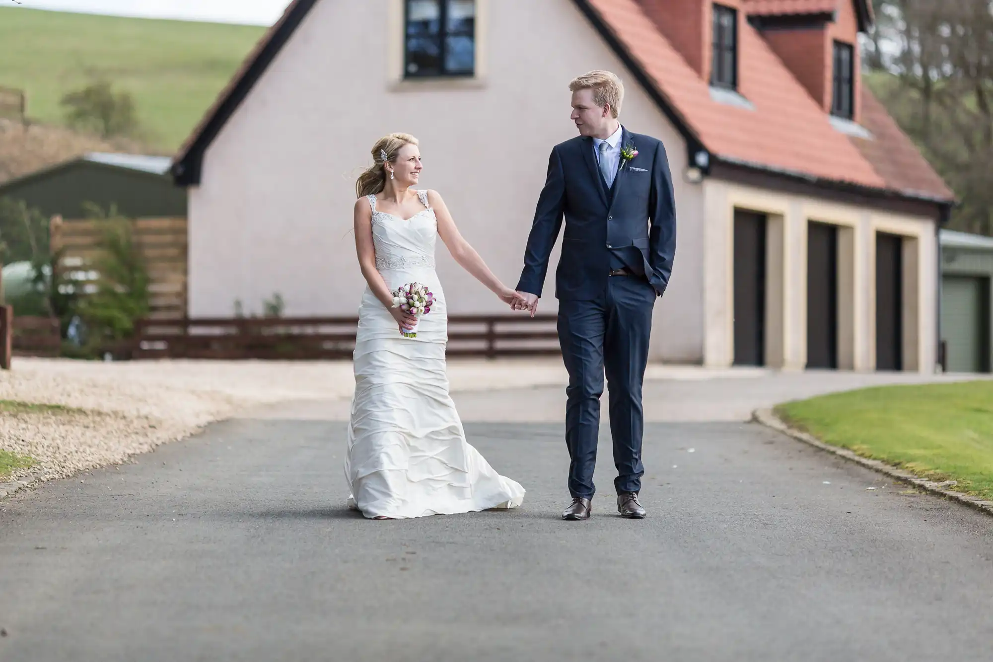 A bride and groom holding hands walk down a paved path, with a house and green hills in the background.