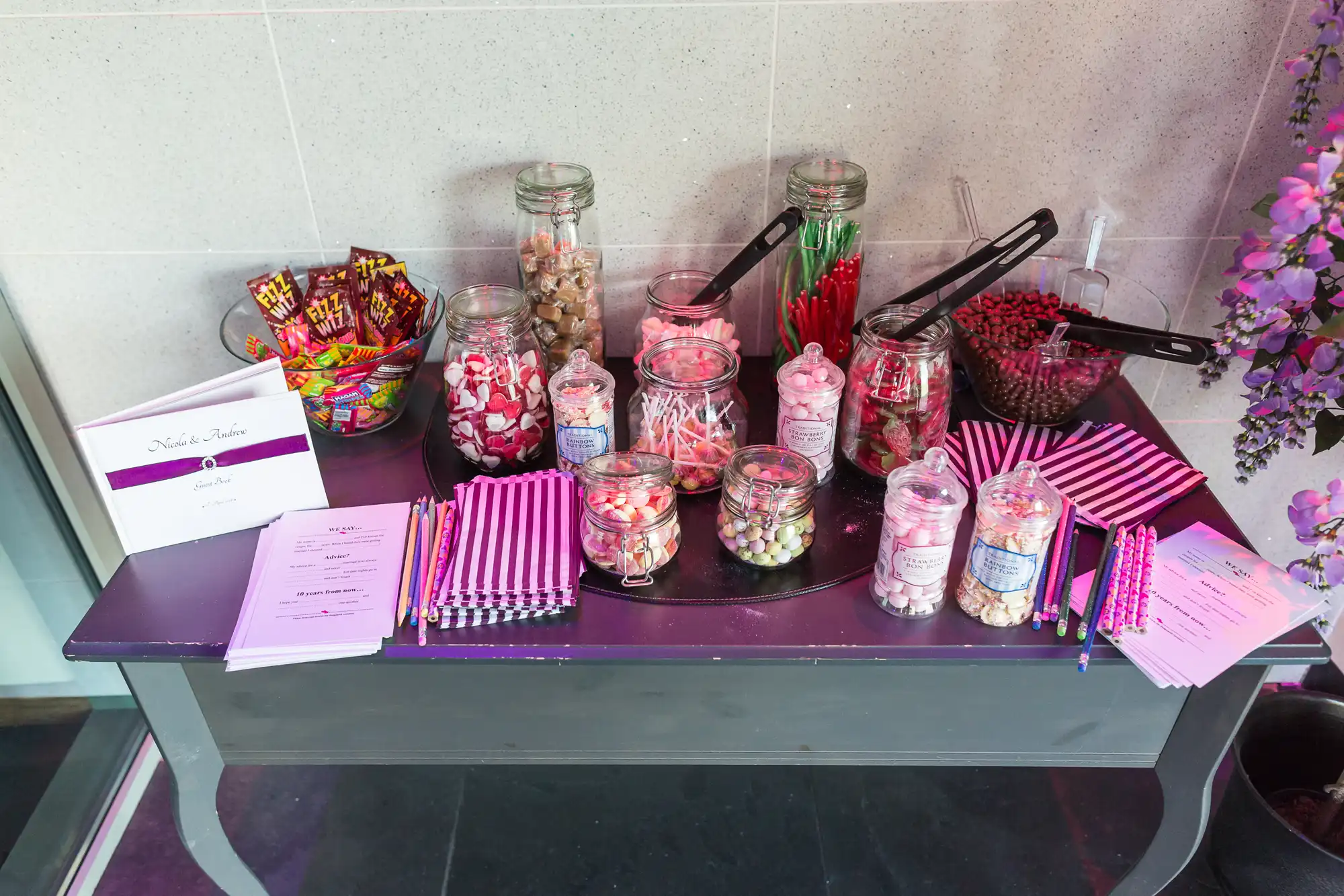 Candy station with various sweets in glass jars and striped paper bags, accompanied by serving utensils and instruction cards, on a dark table.