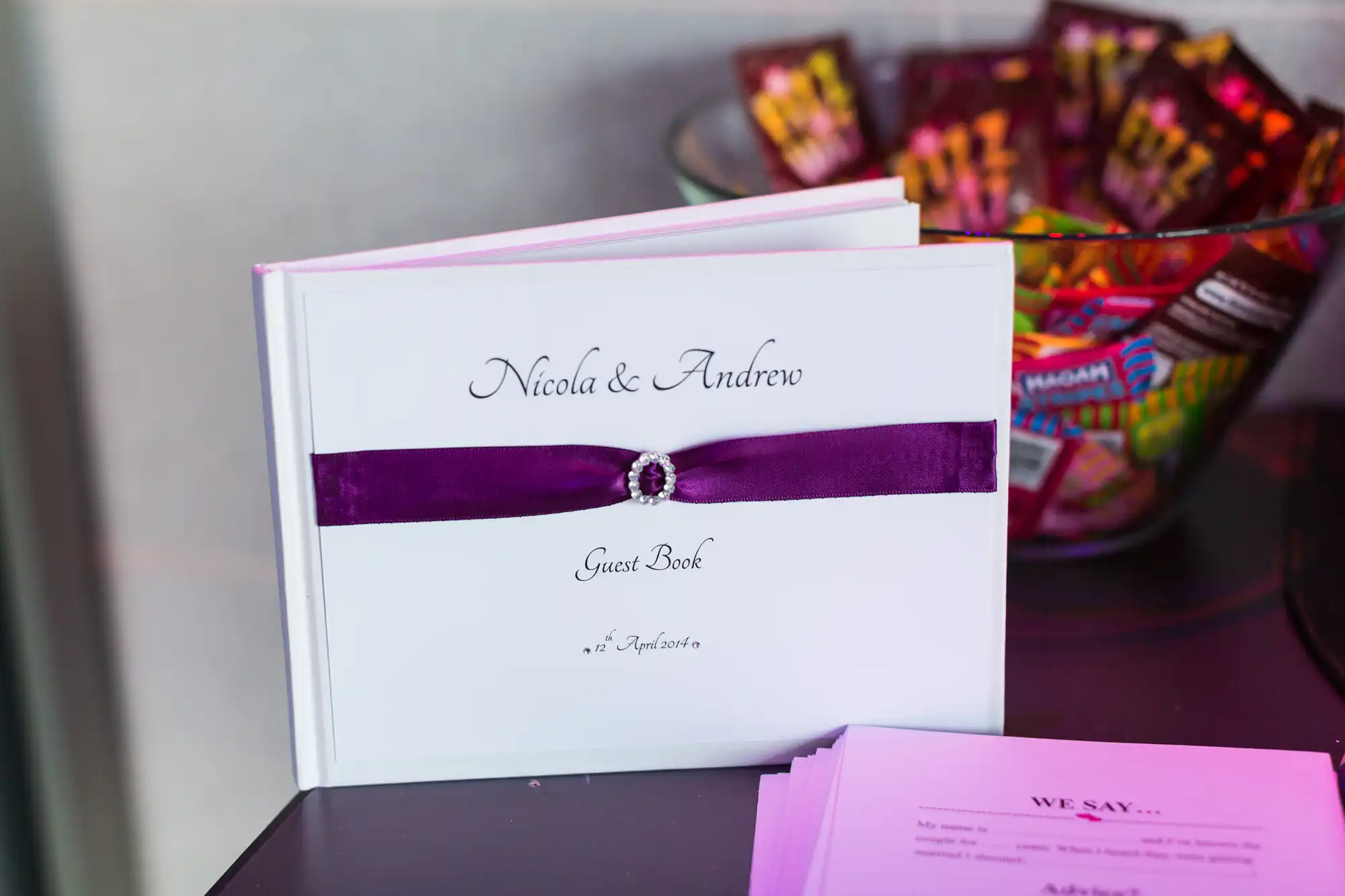 A guest book with "nicola & andrew" in purple text, a purple band and a faux diamond decoration, next to a bowl of wrapped candies.