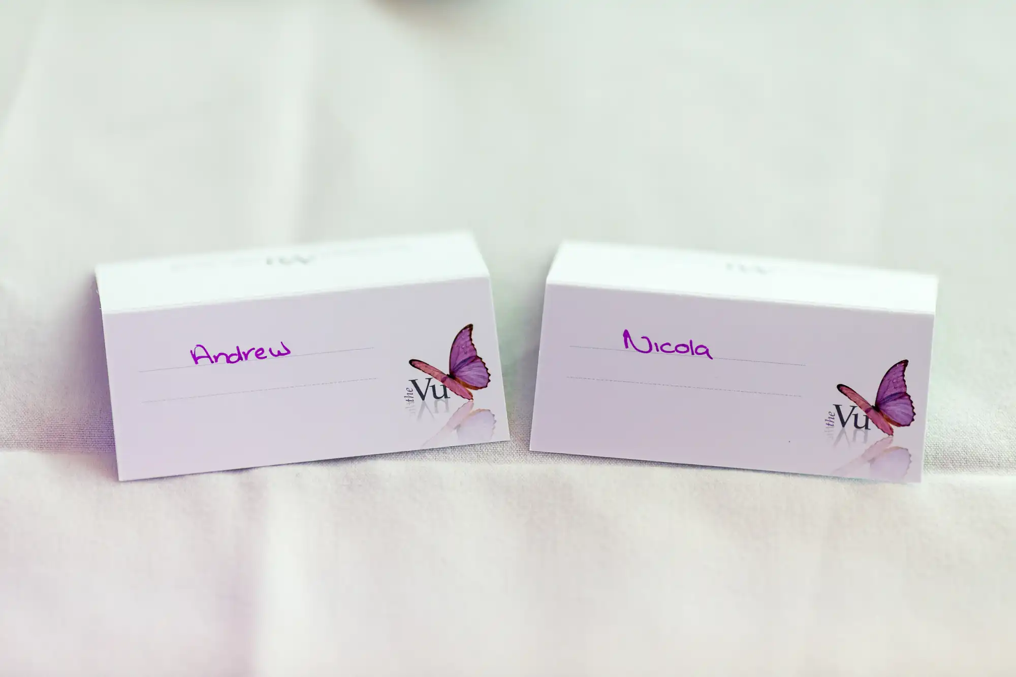 Two place cards reading "andrew" and "nicola" with purple butterfly designs, set on a white linen tablecloth.