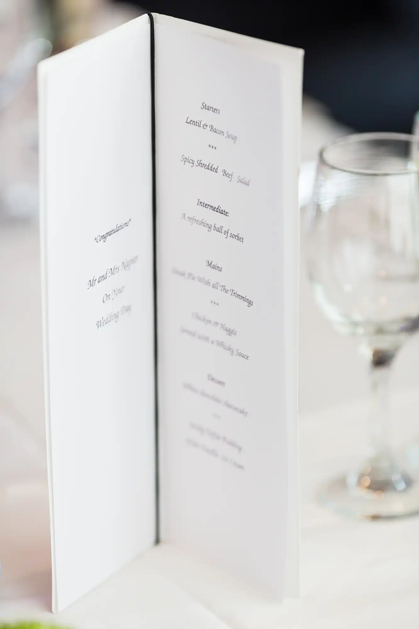 Open wedding menu displaying courses on a table with a blurred glass in the background.