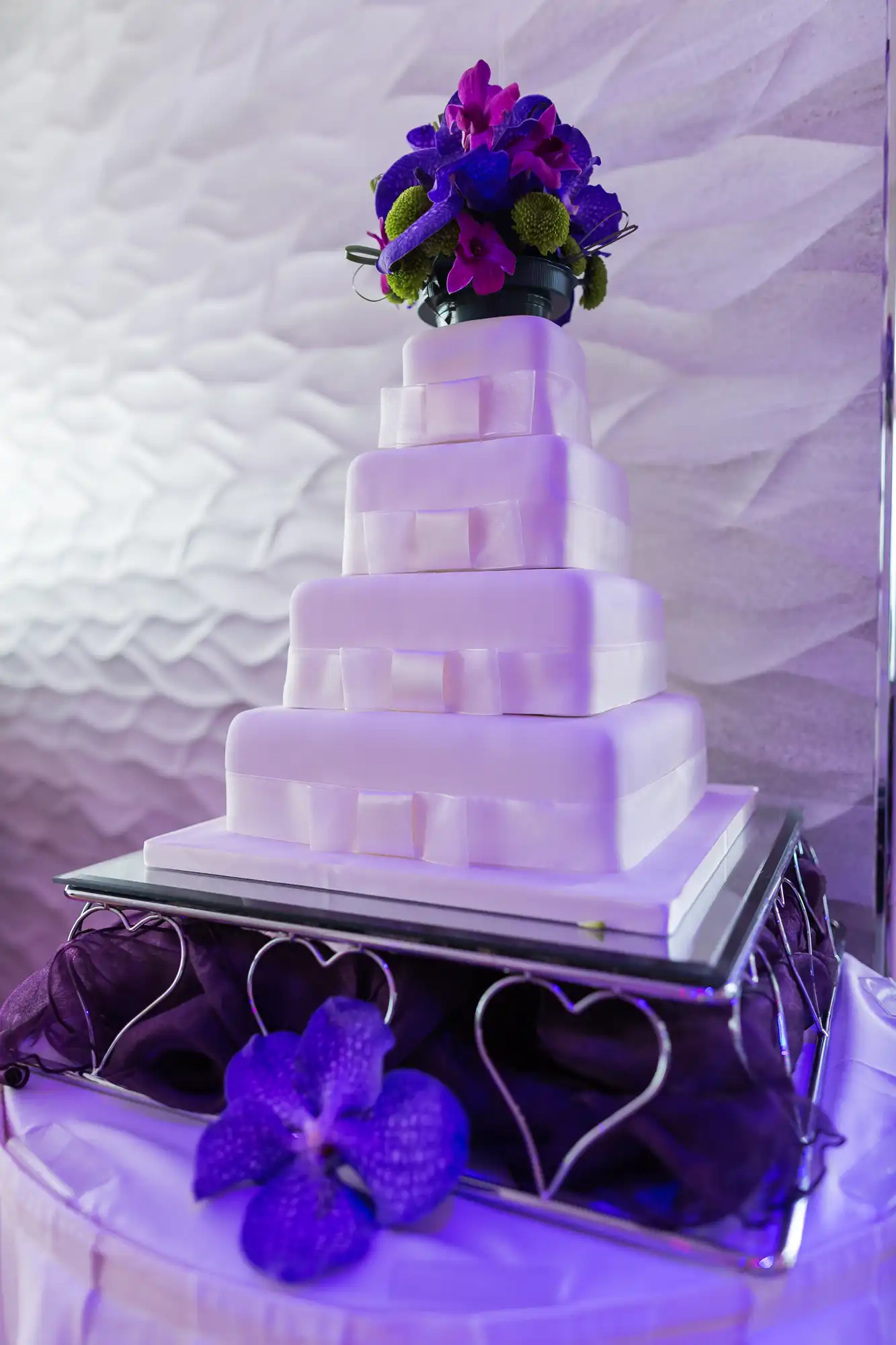 A tiered white wedding cake adorned with purple and blue flowers, displayed on a table with a violet uplighting.