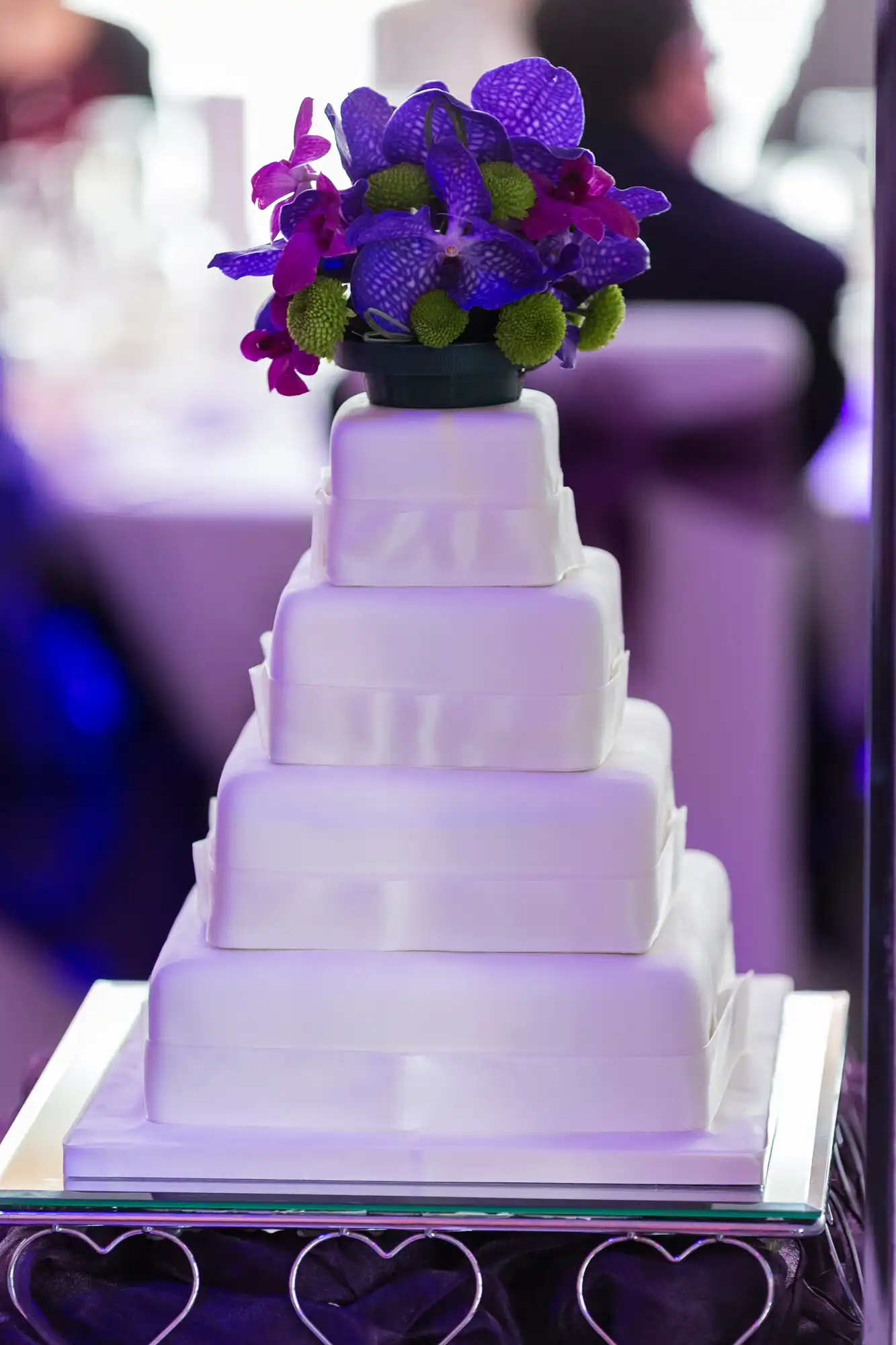 A white multi-tiered wedding cake topped with a bouquet of purple orchids and green blooms, displayed at an indoor event.