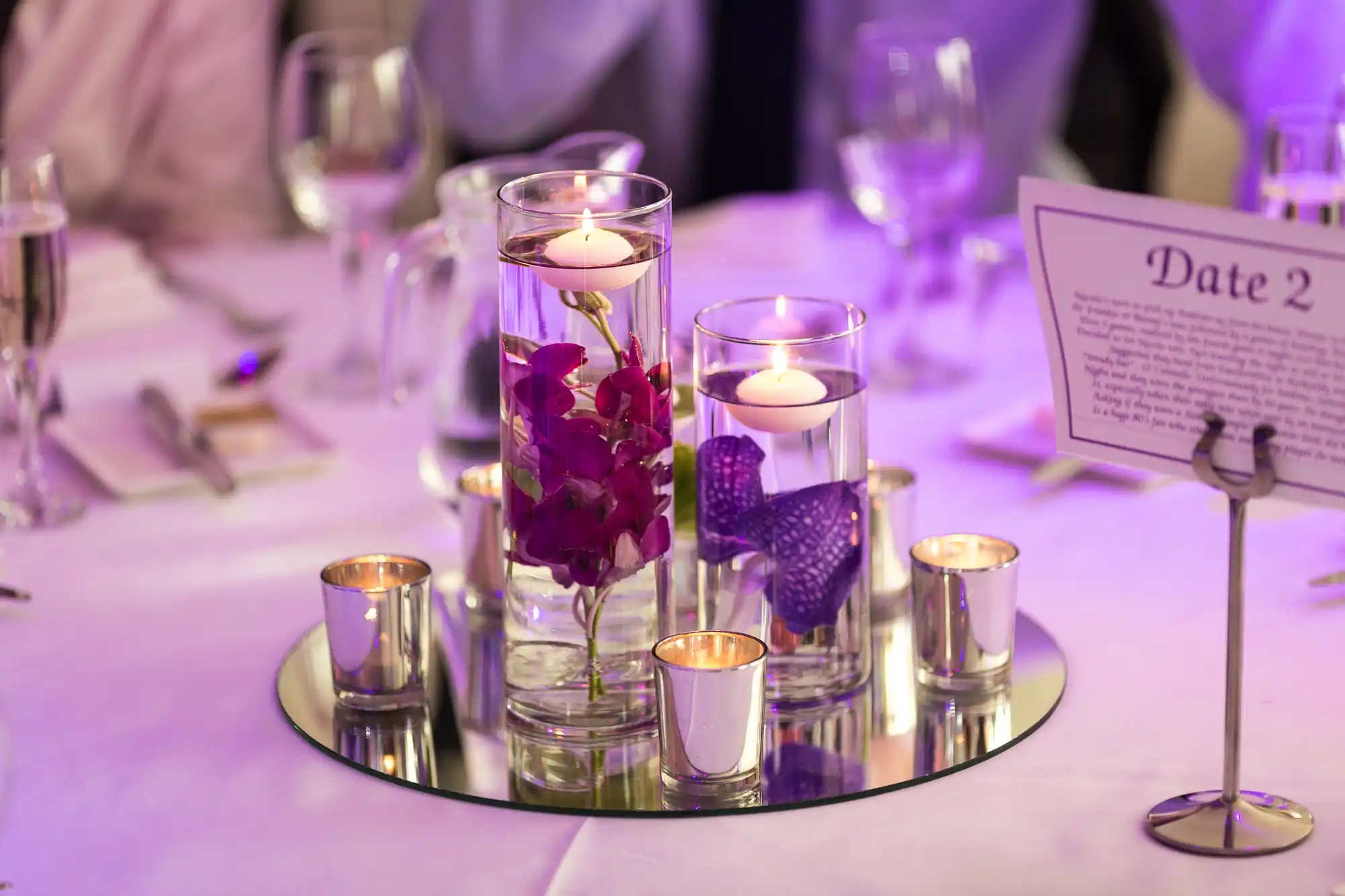 Elegant table centerpiece featuring floating candles in tall glasses, purple flowers submerged in water, and small votive candles, on a reflective mirror base at a formal event.