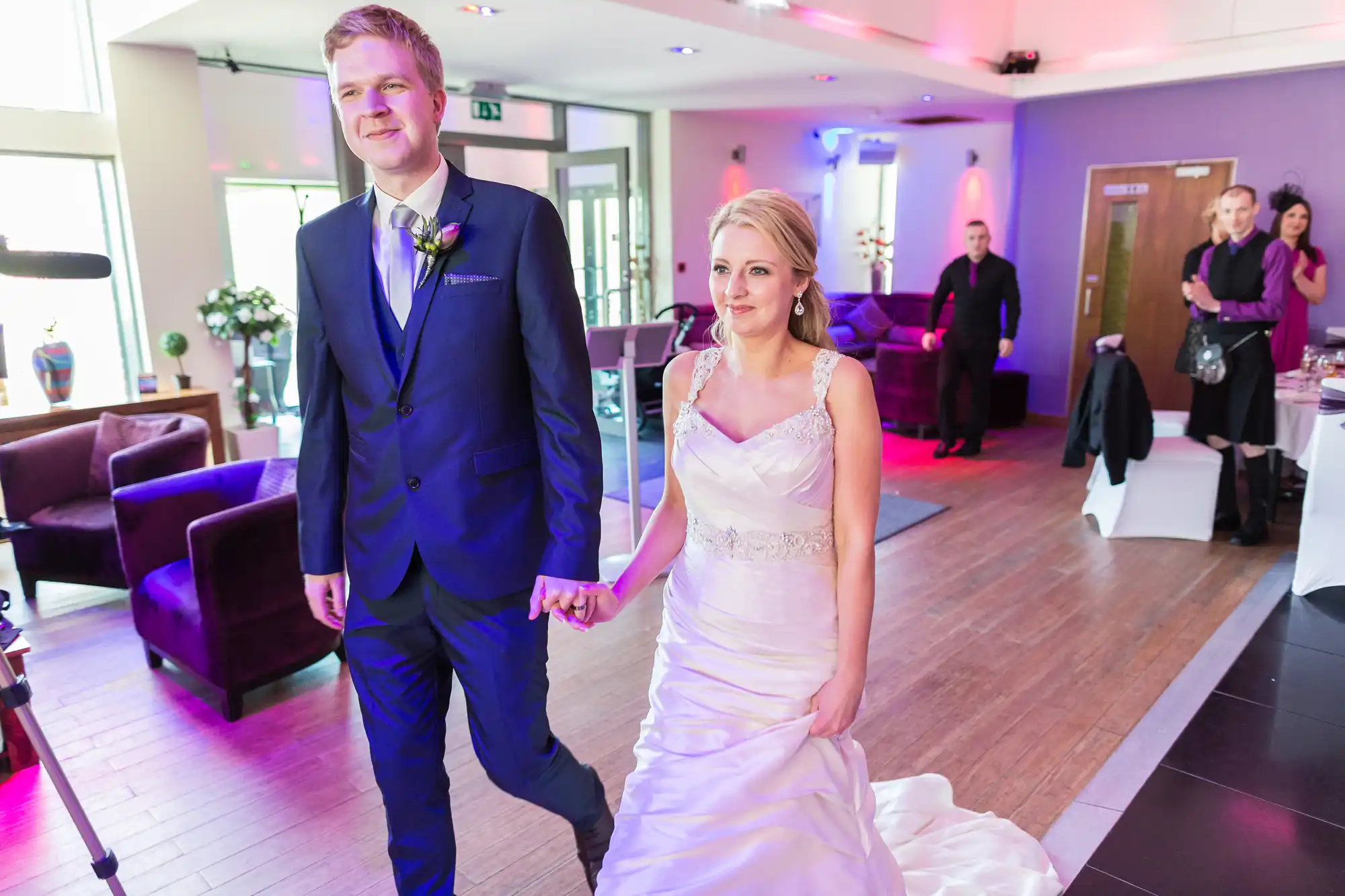 A bride and groom holding hands and walking through a modern venue, with guests in the background.