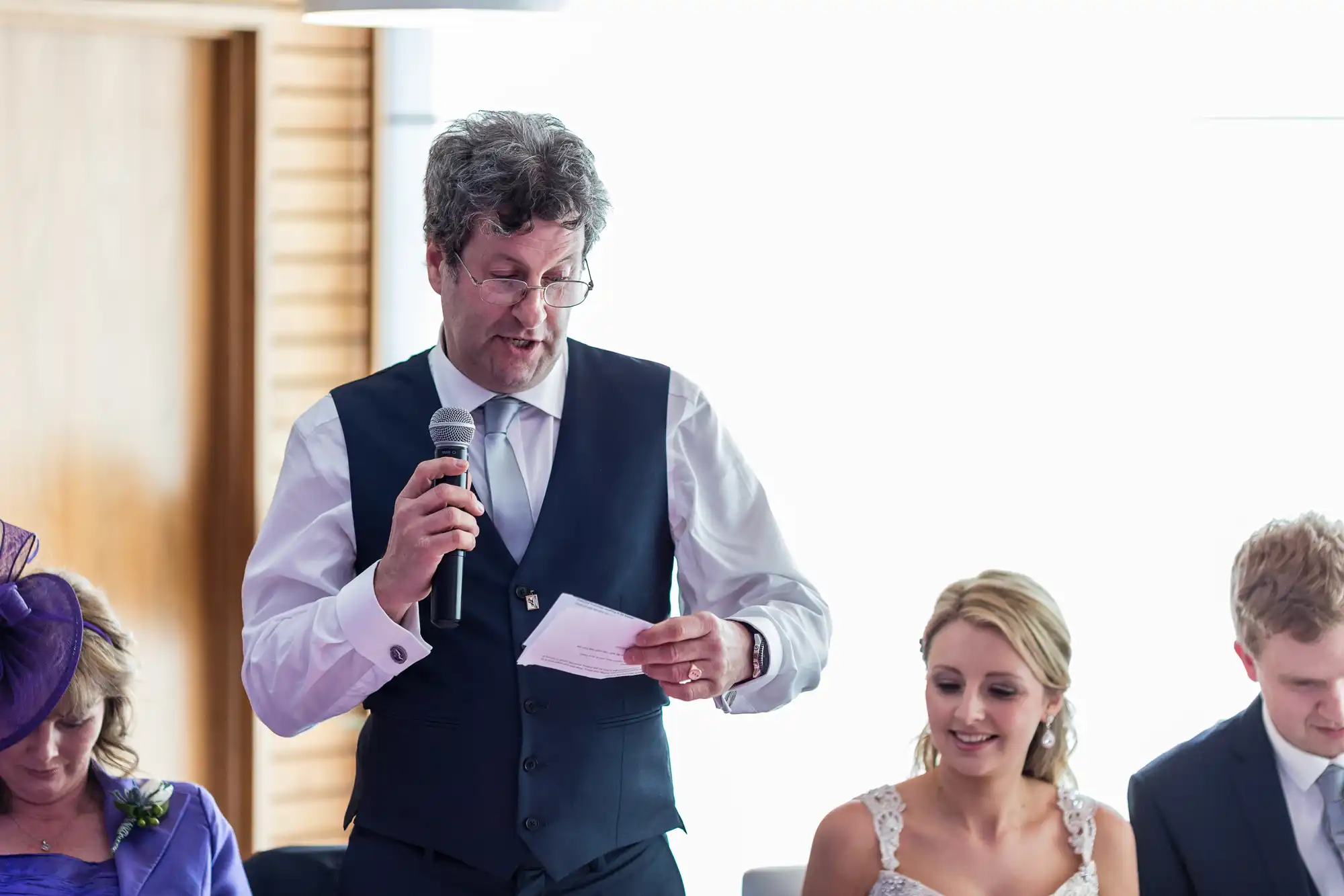 A man in a vest and tie delivers a speech with a microphone at a formal event, holding a paper, with seated guests listening attentively.