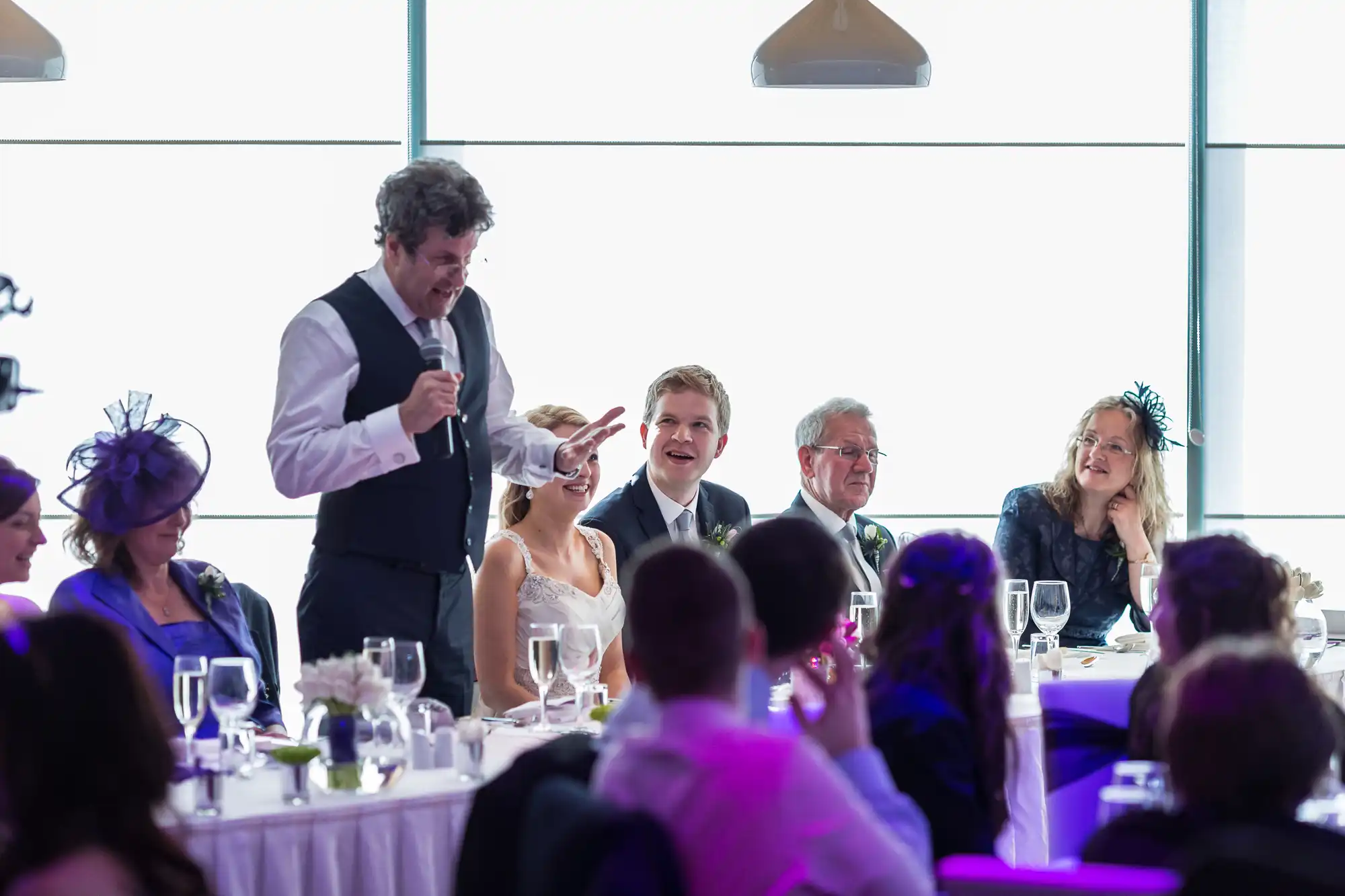 A man gives a speech at a wedding reception, standing beside a bride and groom at a table, with guests listening attentively.