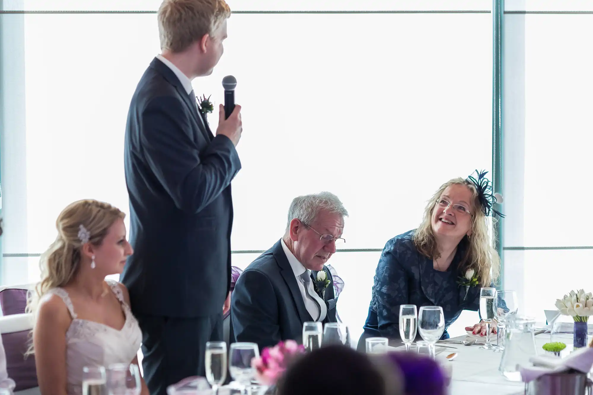 A man standing and speaking into a microphone at a wedding reception, with a laughing couple and a woman listening at a table, in a room with large windows.