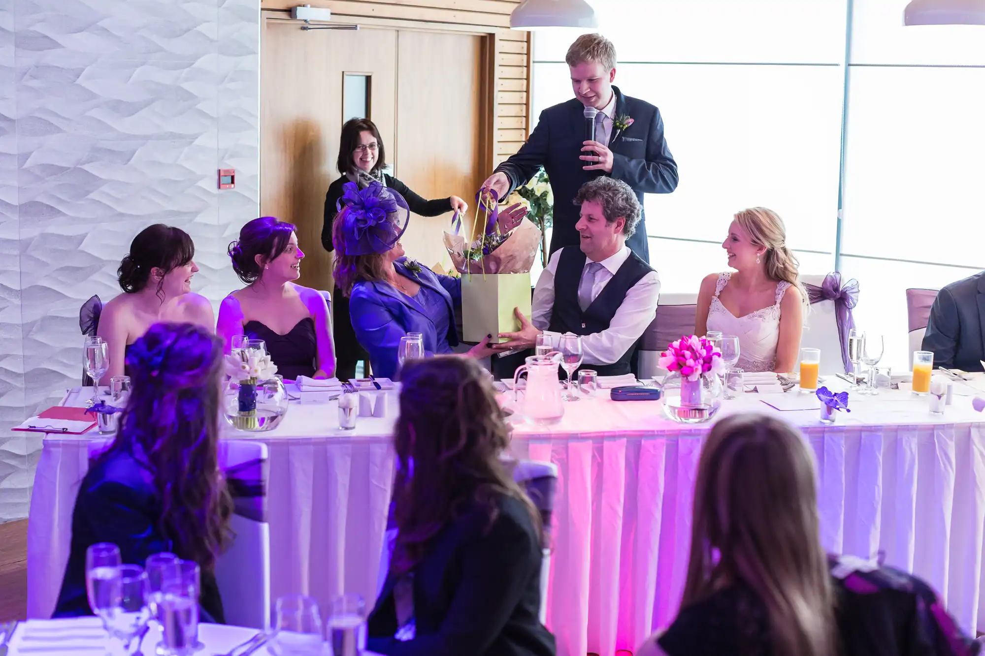A wedding reception scene with guests seated at tables, focusing on a standing man giving a speech to a happy couple.