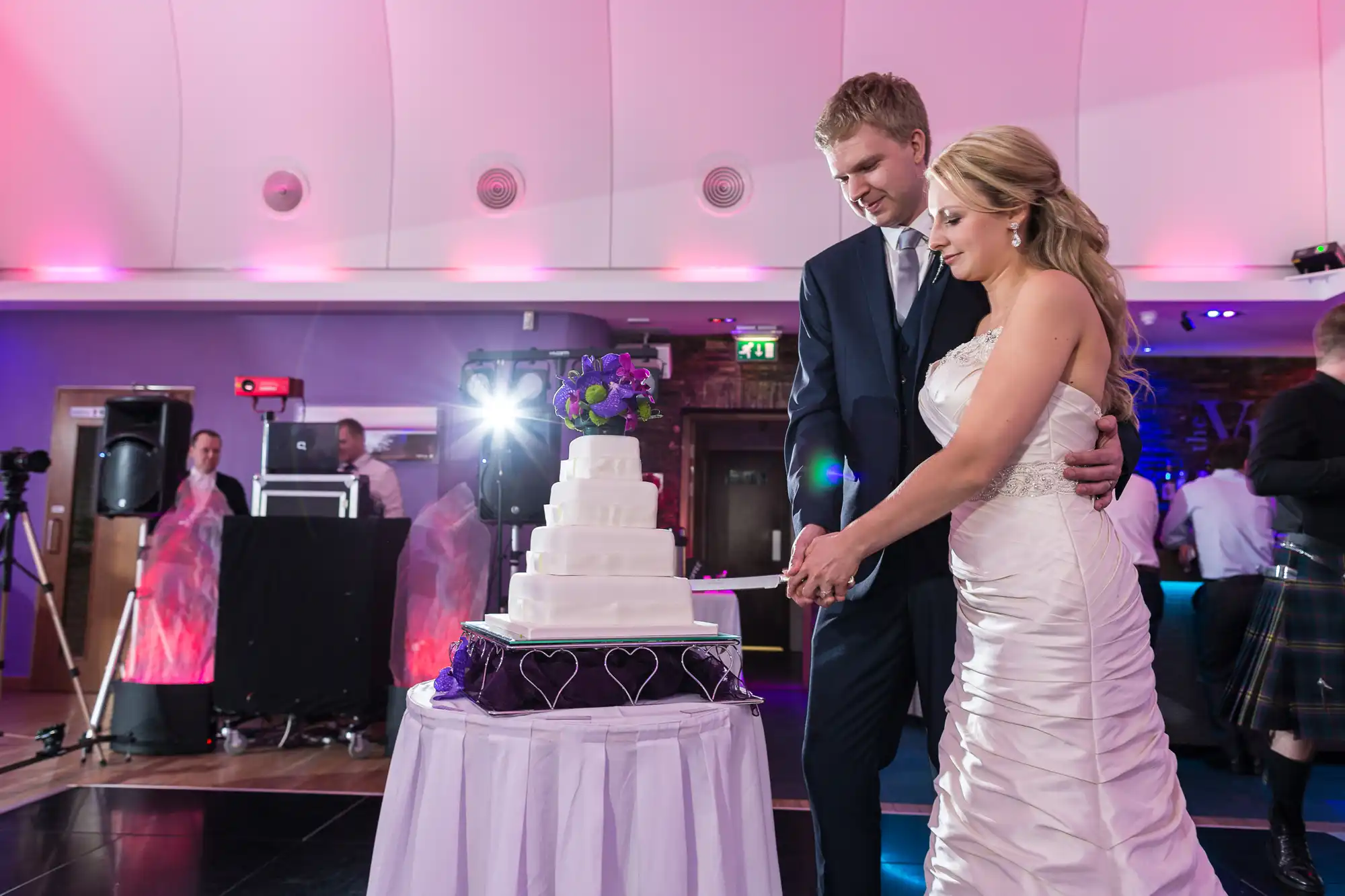 A bride and groom cutting a multi-tiered wedding cake in a banquet hall with purple lighting and a dj setup in the background.