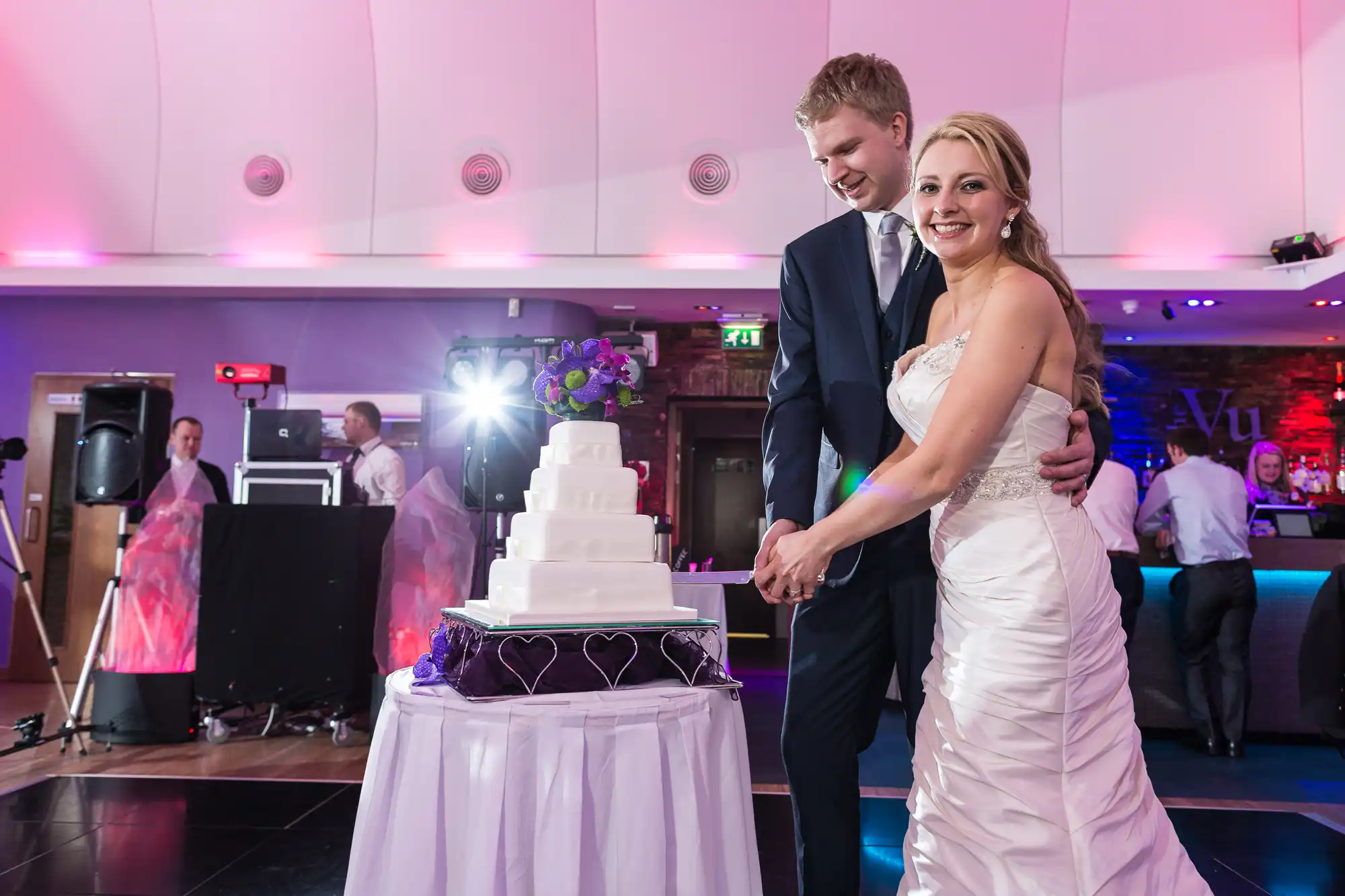 A bride and groom smiling while cutting a multi-tiered wedding cake at a reception hall with purple lighting and a dj booth in the background.