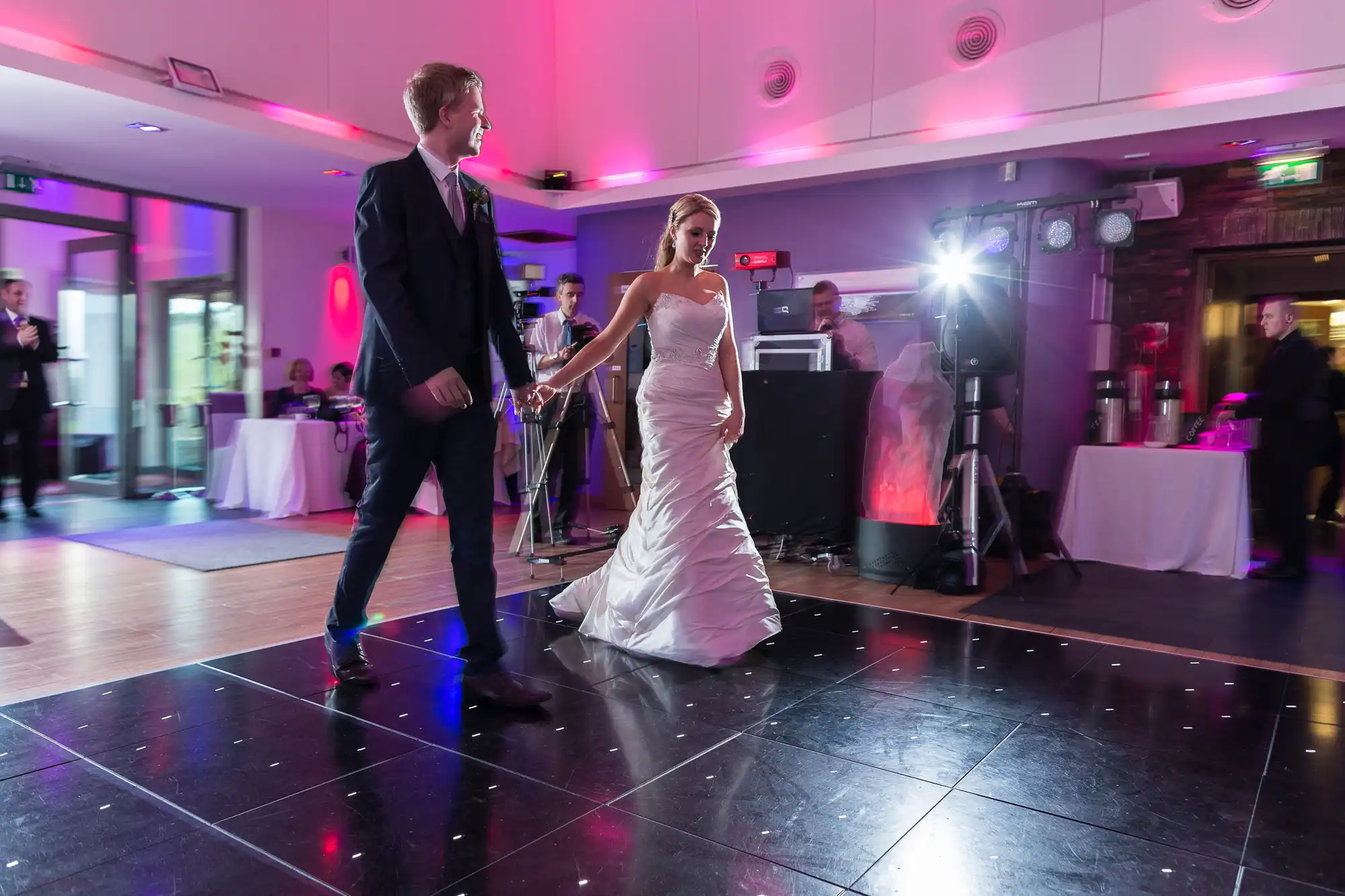 A bride and groom holding hands and dancing at their wedding reception, surrounded by guests and colorful lighting.