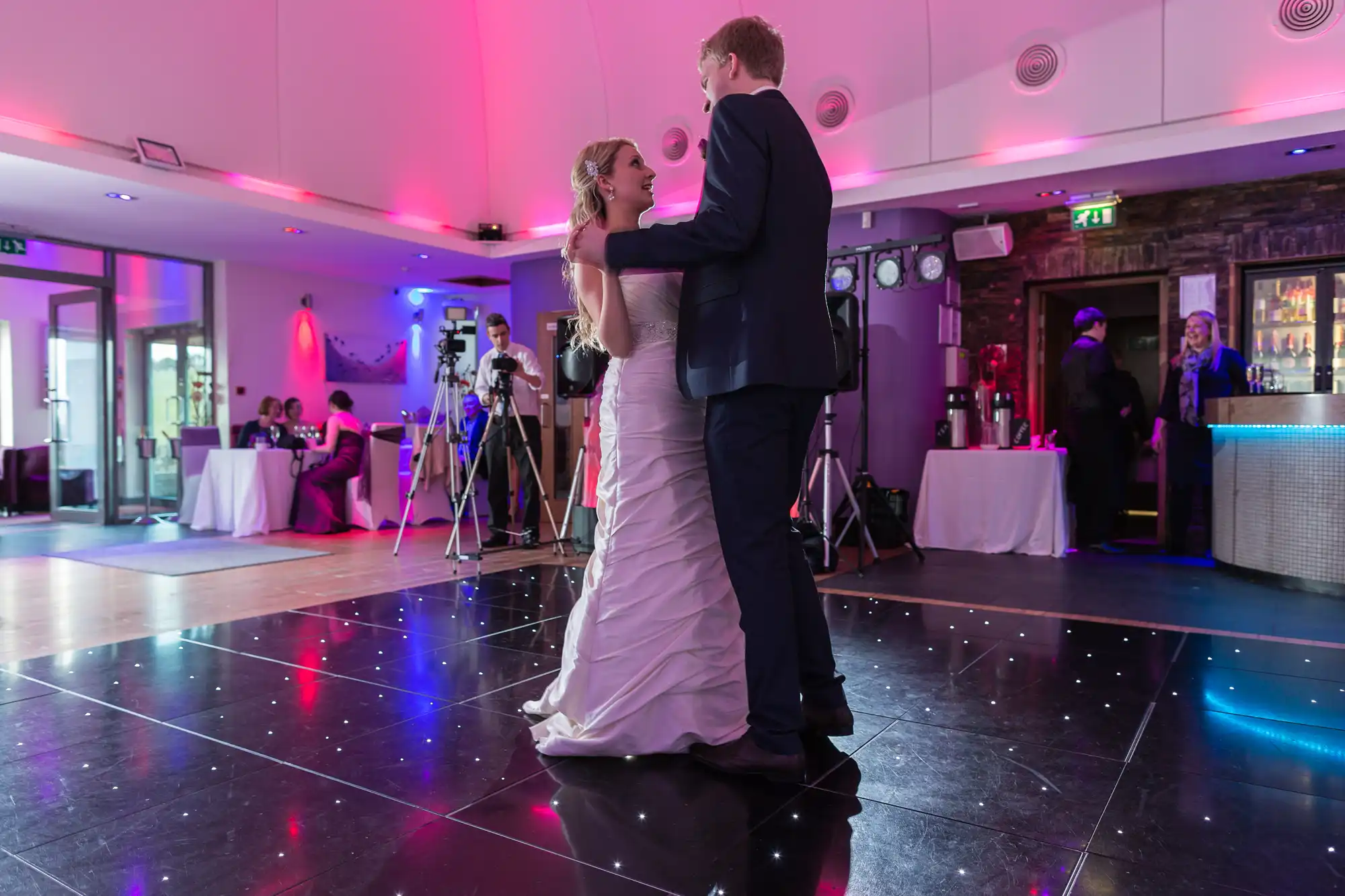 A bride and groom share a dance in a dimly lit venue with pink lighting, surrounded by guests and a video camera on a tripod.