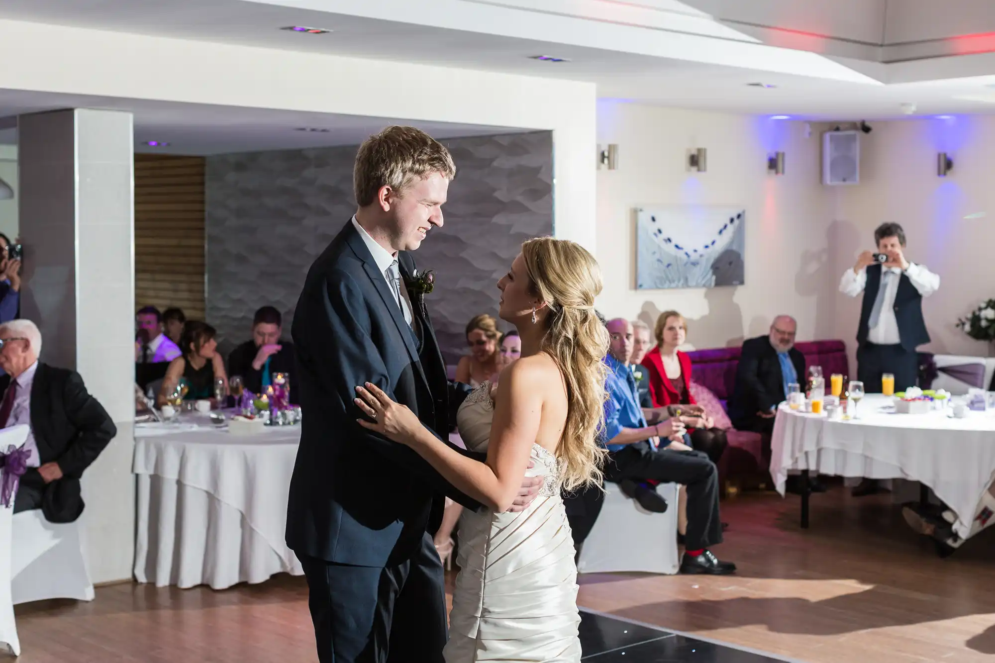 A newlywed couple sharing their first dance in a banquet hall, surrounded by seated guests, with soft lighting and a person capturing the moment.