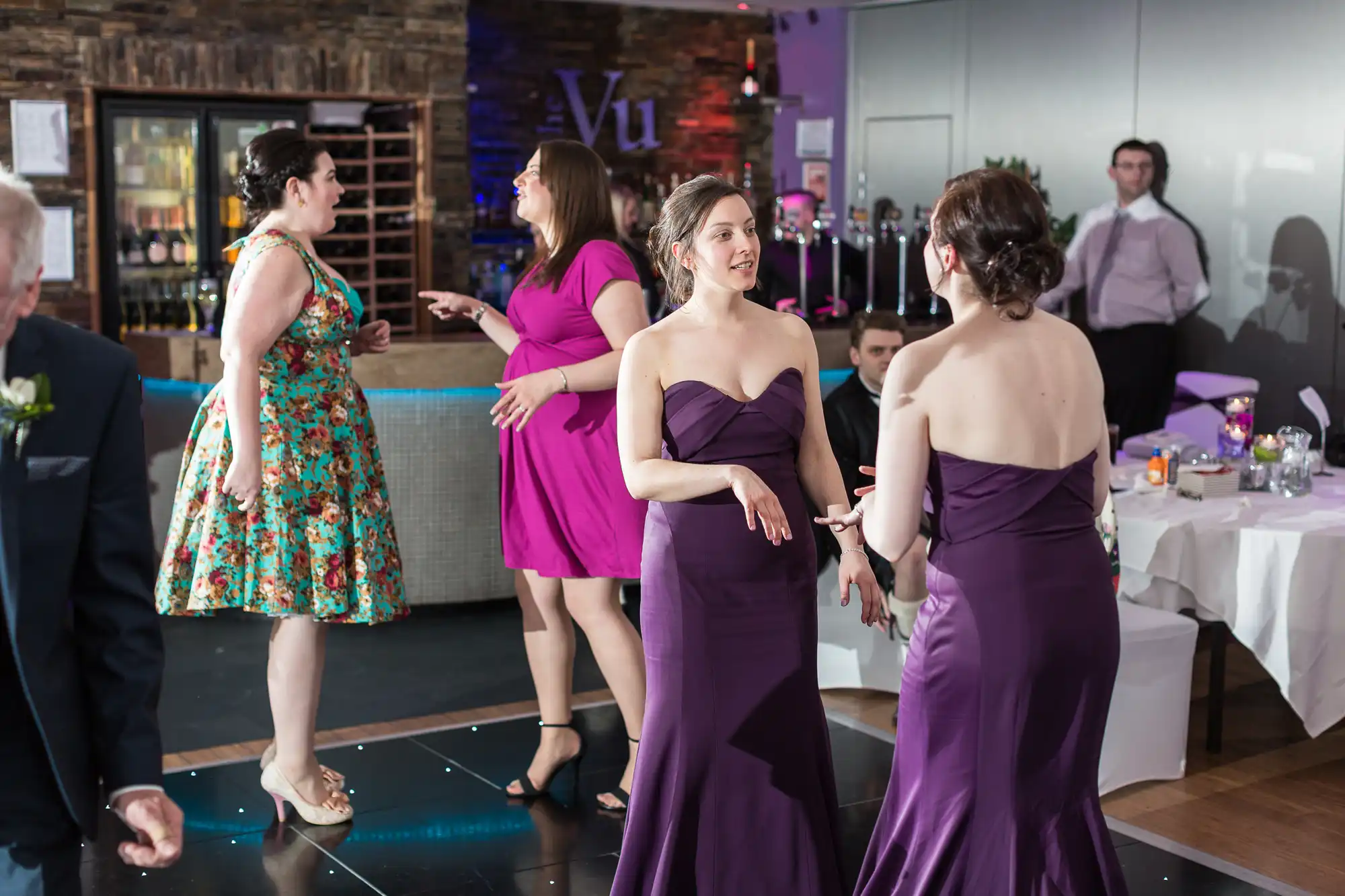 Women in formal attire dancing and conversing at an indoor celebration, with tables and party decorations visible in the background.