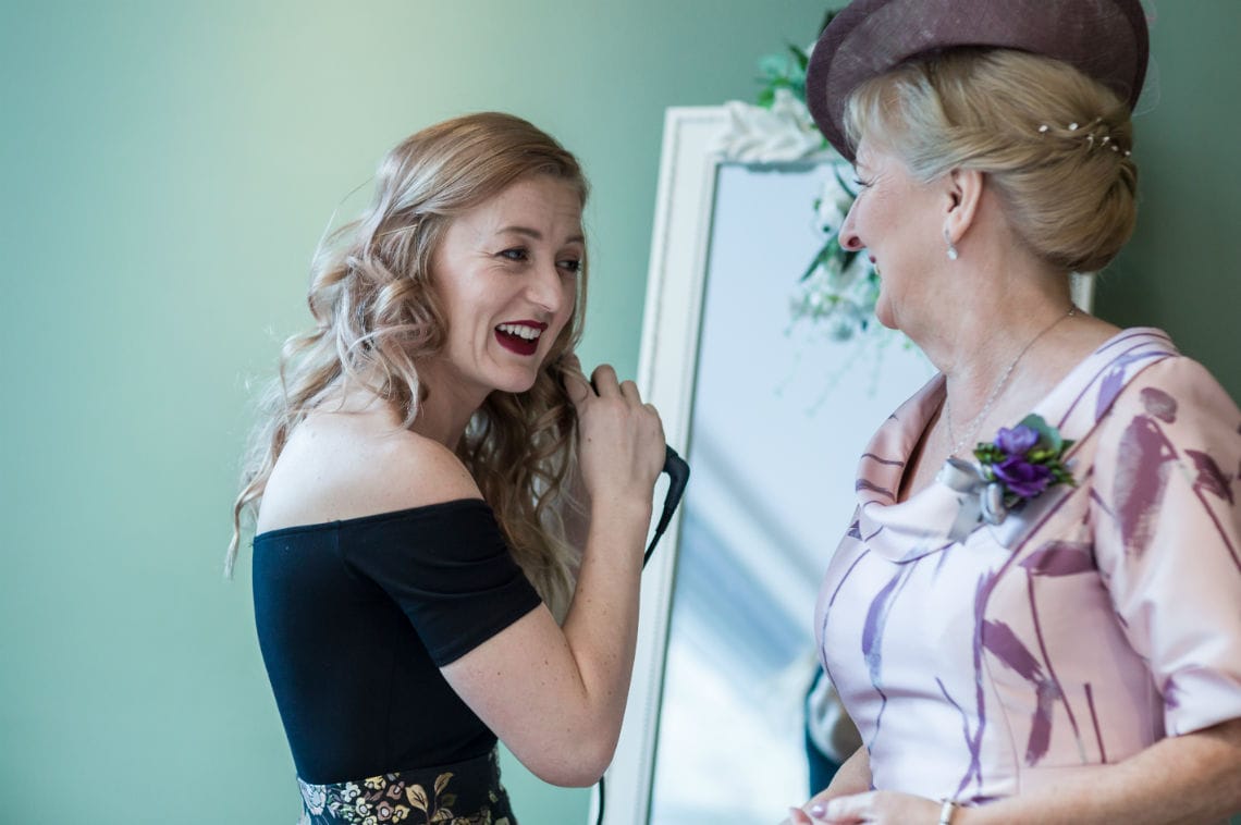 lady smiling at mother of the groom as she curls her hair in front of mirror during bridal preparations