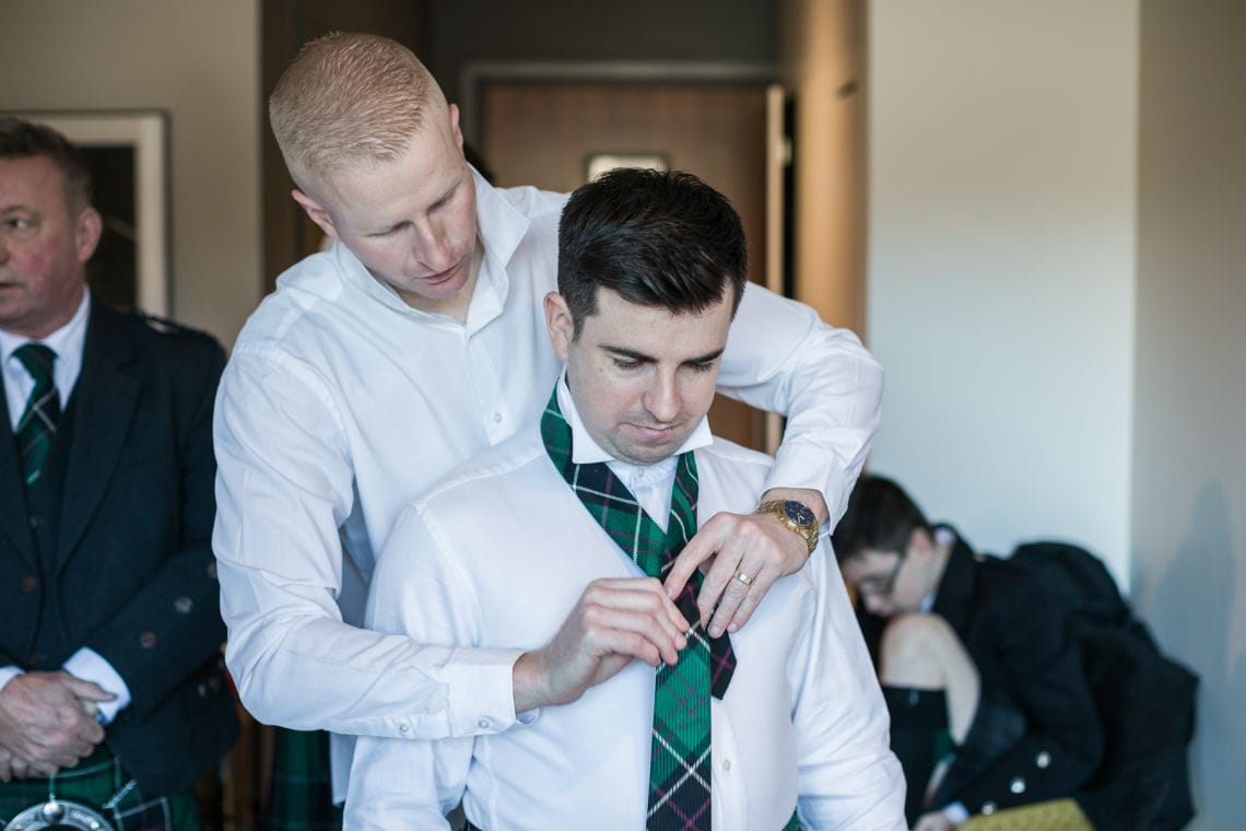 Guest wearing white shirt standing behind groom putting green tartan tie on groom