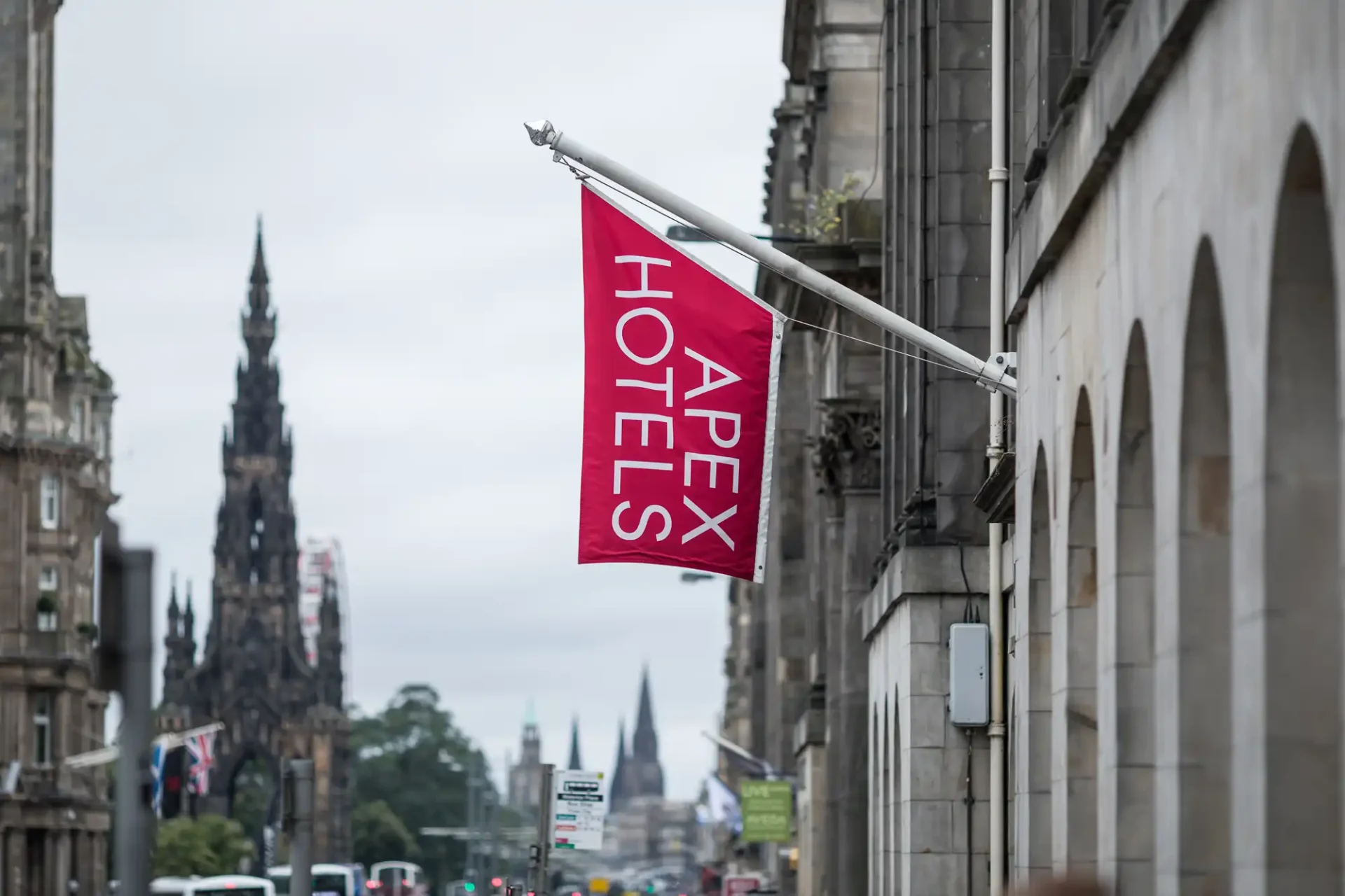 An apex hotels flag protrudes from a building, with edinburgh's scott monument visible in the background under a cloudy sky.