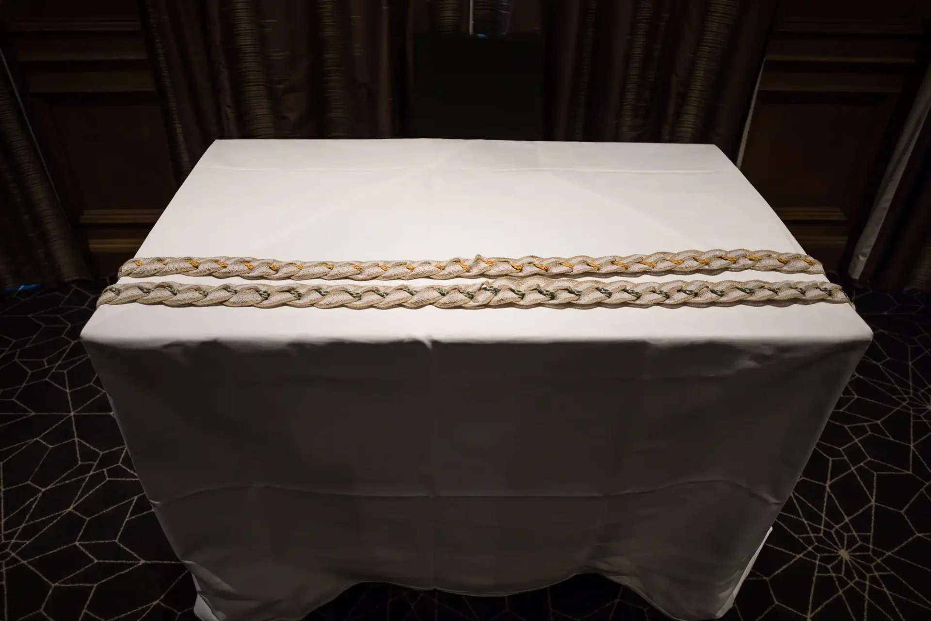 A neatly draped white tablecloth on a rectangular table, accented with an intricate golden braid along the front edge.