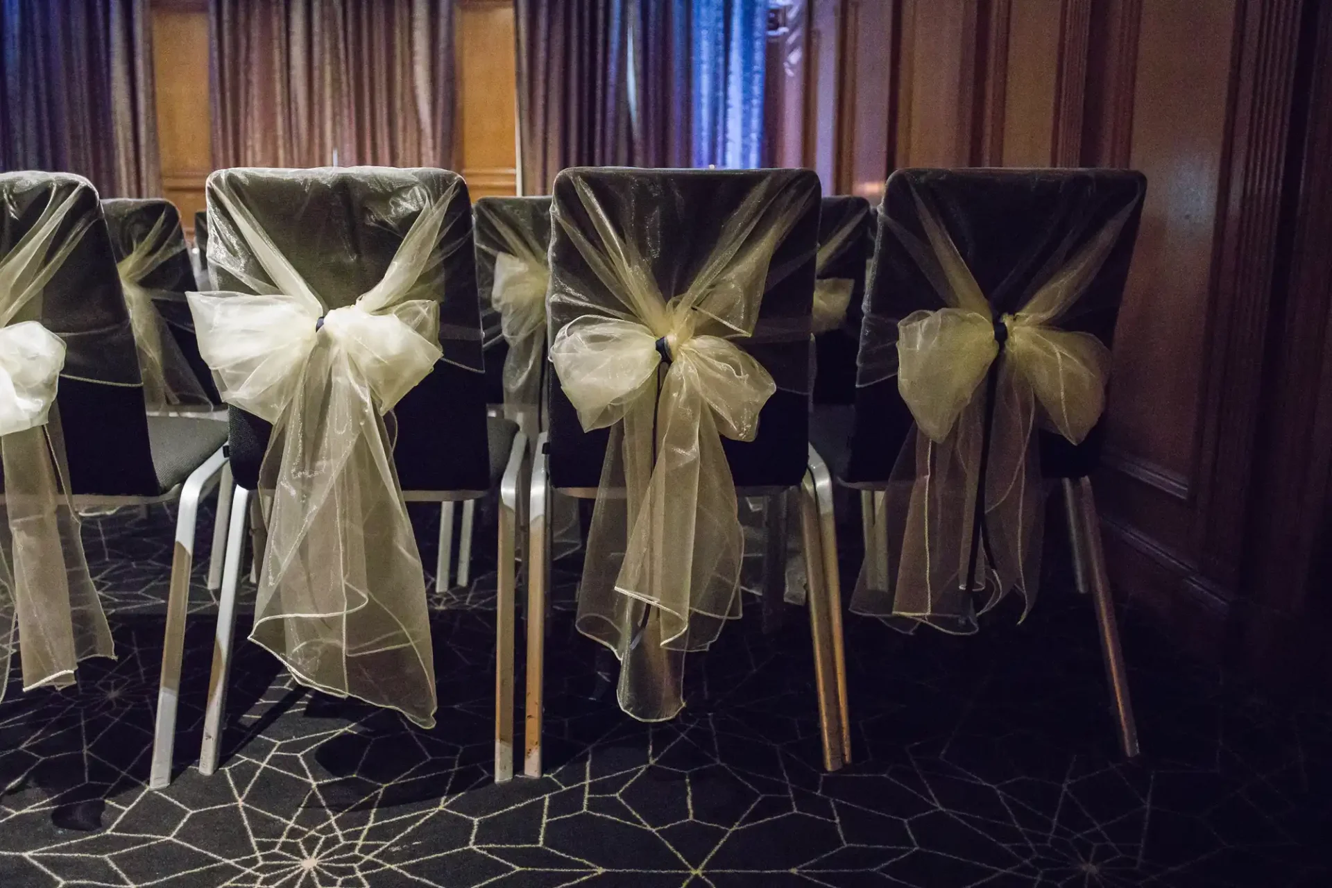 Rows of chairs covered with black fabric and decorated with white sheer bows in a dimly lit room.