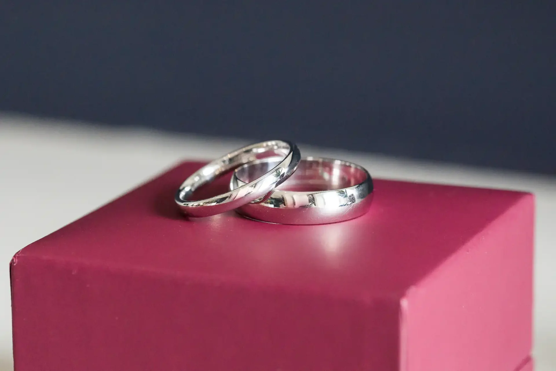 Two wedding rings on a pink box, one ring engraved with a visible design, against a soft-focus background.