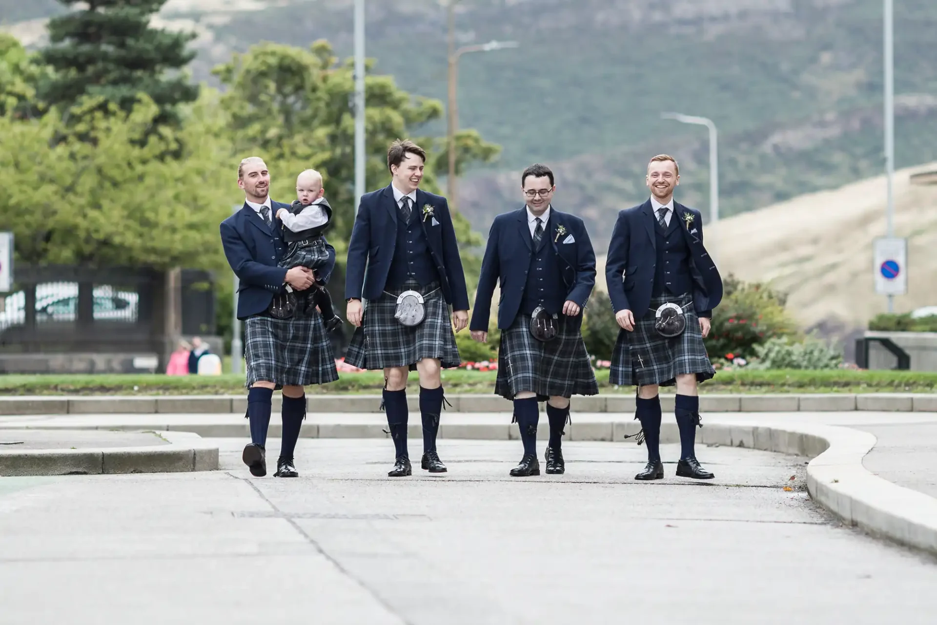 Five men in traditional scottish attire, including kilts, walking together in a city setting, with one holding a young child.