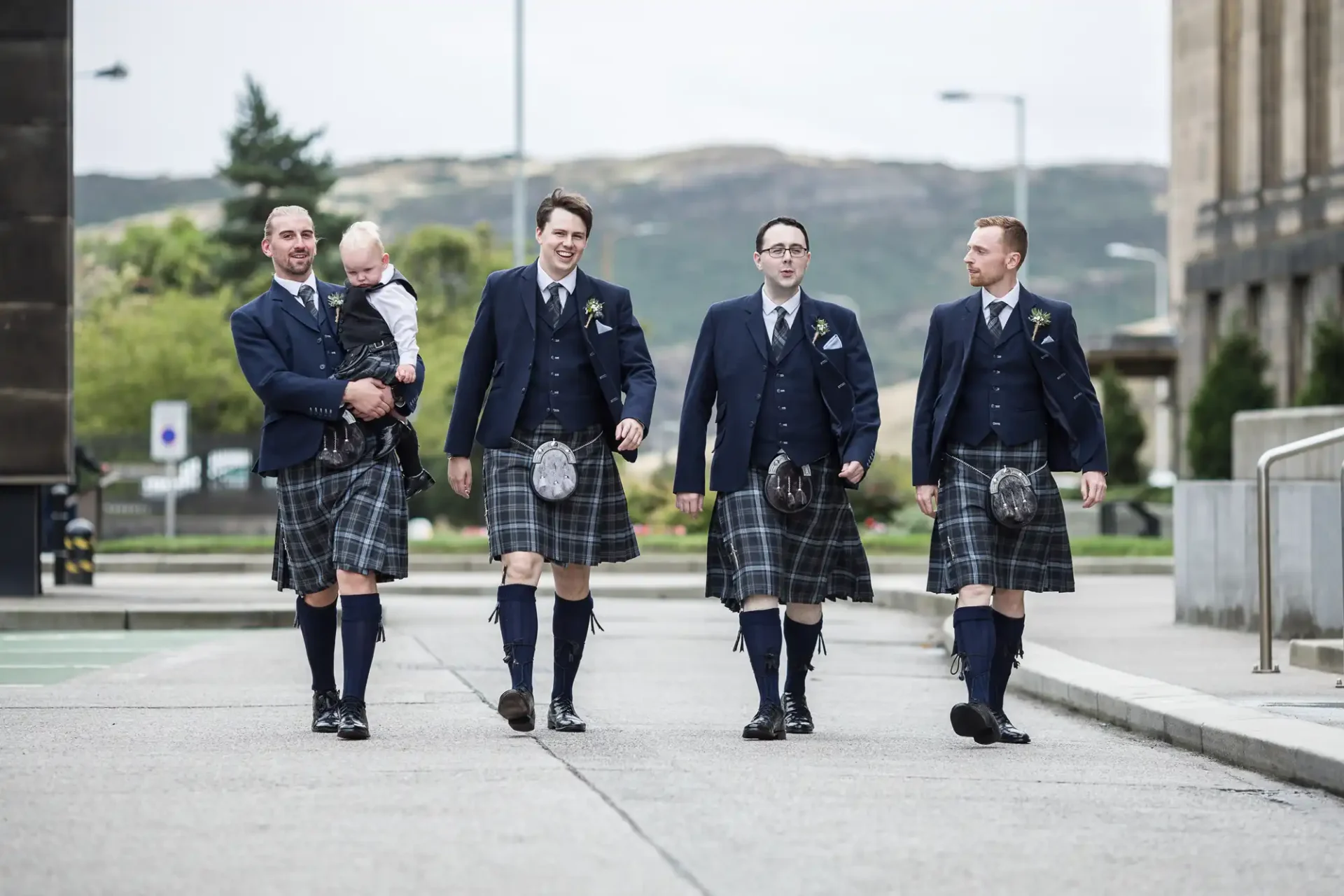 Four men and a baby wearing traditional scottish kilts and jackets walking on a paved path, with urban buildings in the background.