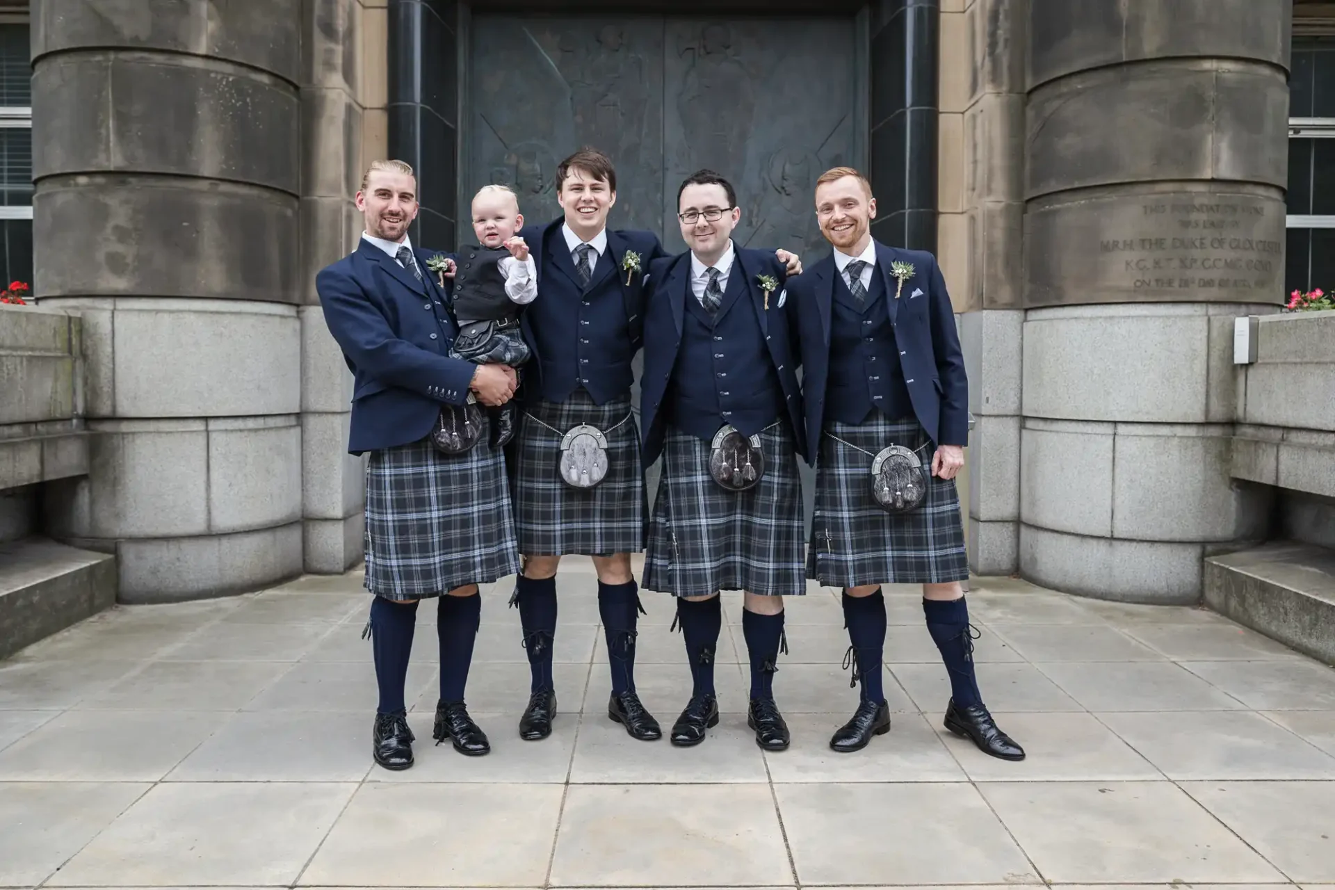 Four men and a baby, all in matching tartan kilts and jackets, smiling outside a stone building.
