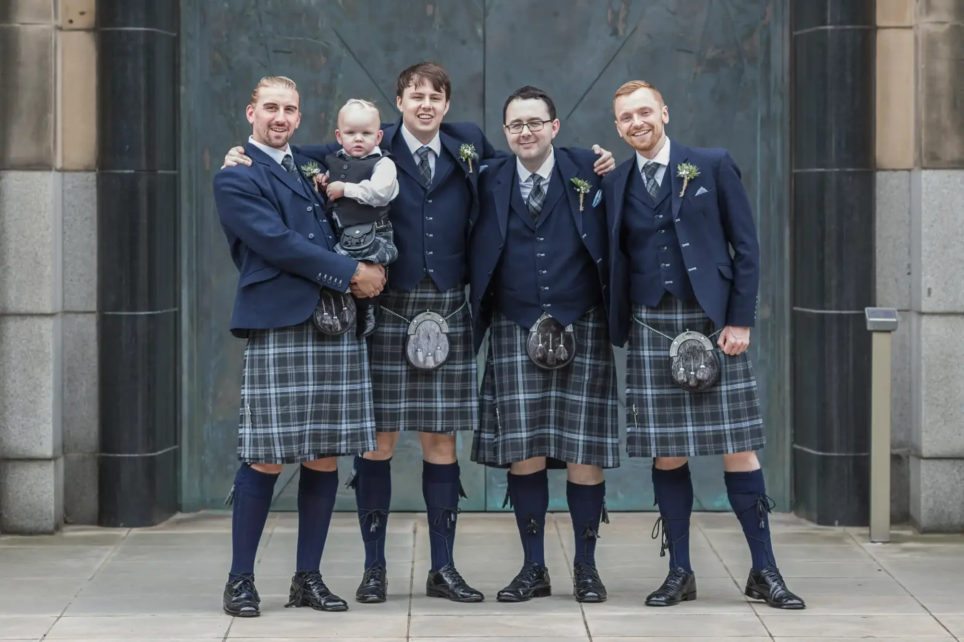Four men in traditional scottish kilts and jackets, smiling with a baby, standing against a stone columned backdrop.