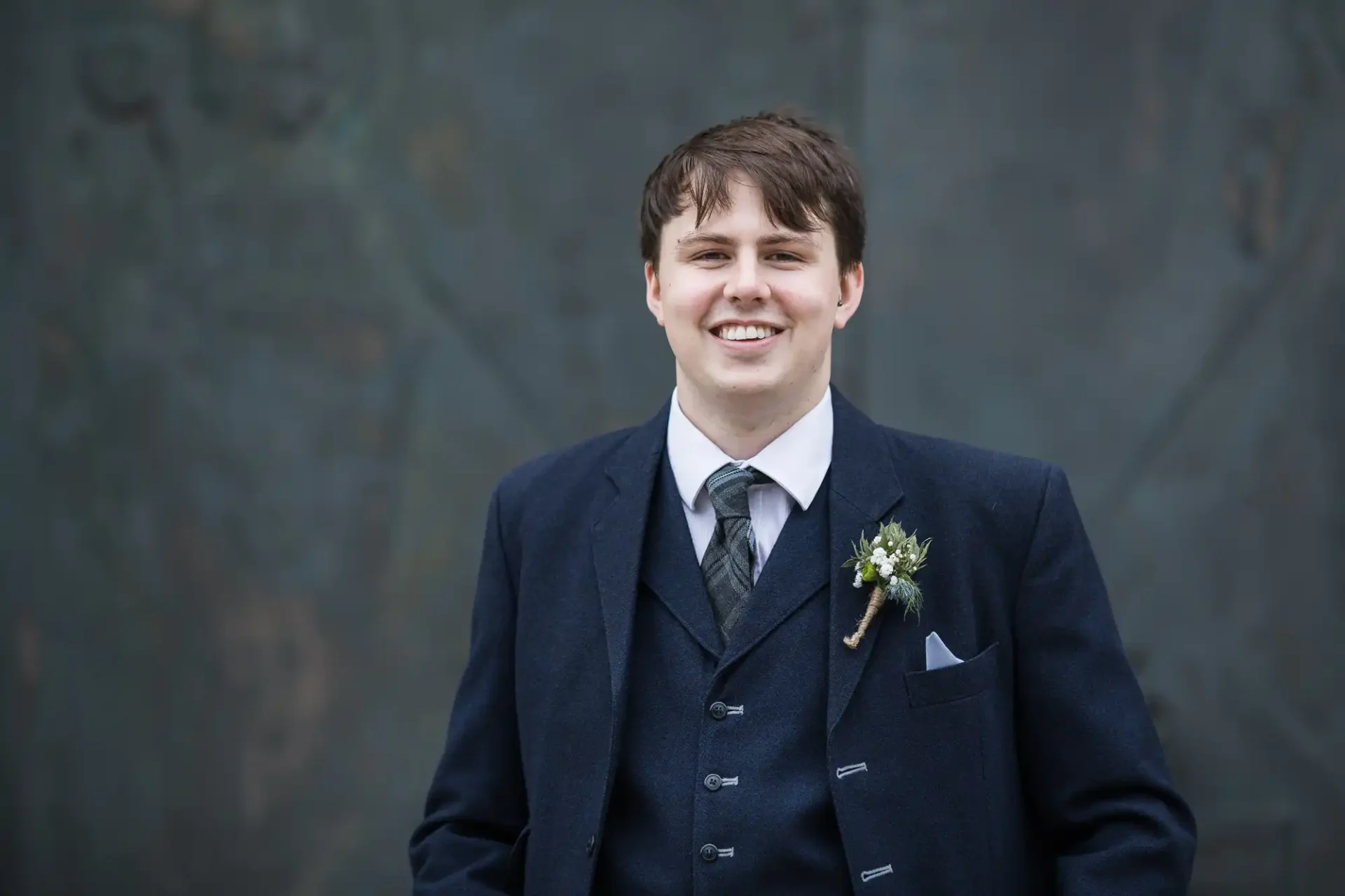A smiling groom in a dark blue suit with a boutonniere, standing in front of a muted, textured backdrop.