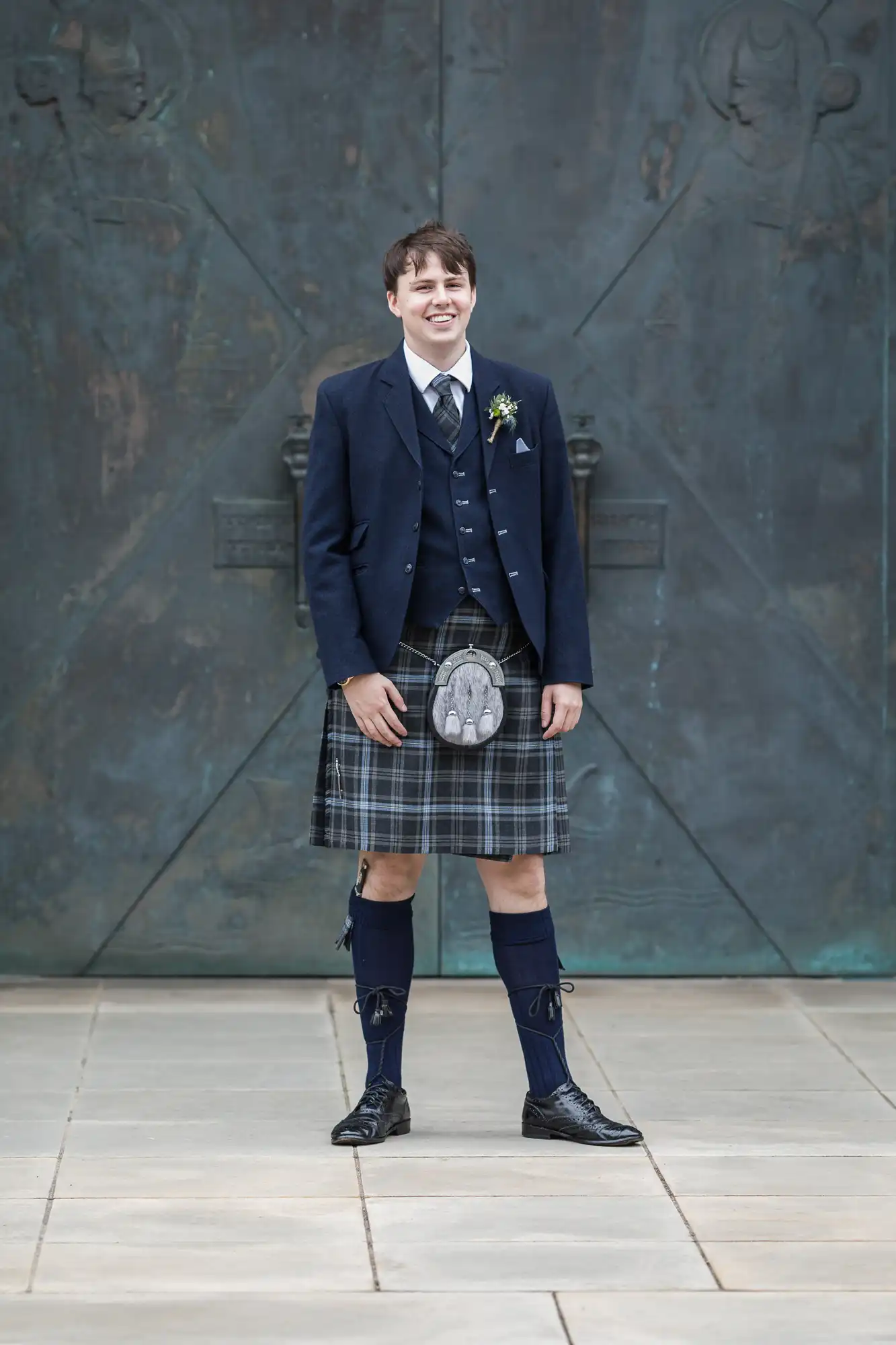 A young man in formal attire with a kilt, smiling and standing before a wall with bas-relief sculptures.