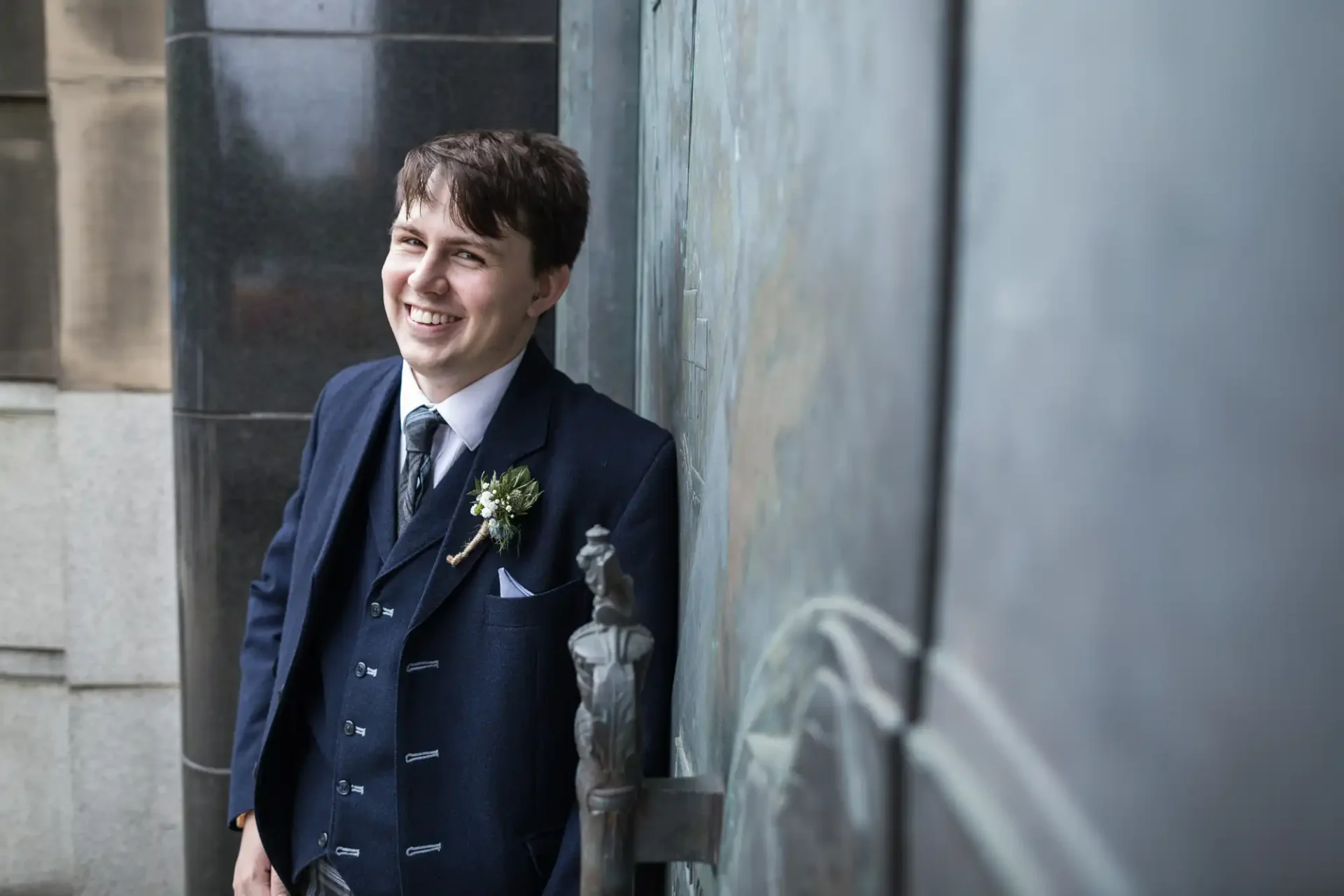 Young man in a dark suit smiling beside an ornate metal doorway, with a flower boutonniere on his lapel.