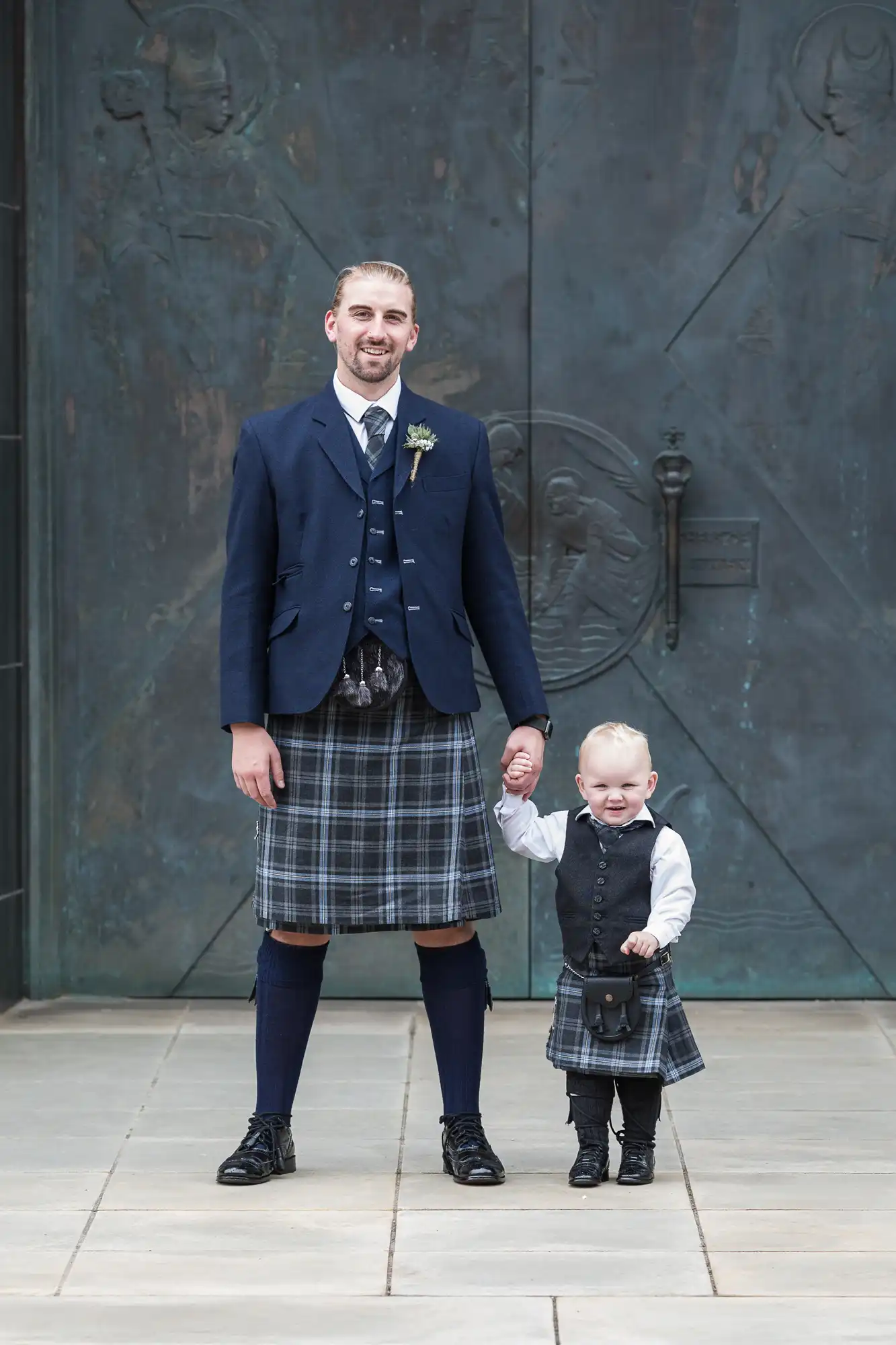 A man in a kilt and jacket stands smiling beside a young child in a matching kilt outfit, both posing in front of a relief sculpture wall.