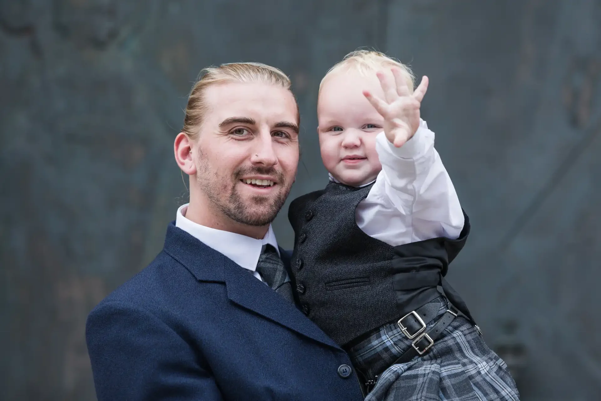 Man in a suit holding a toddler in a kilt, who is waving at the camera.