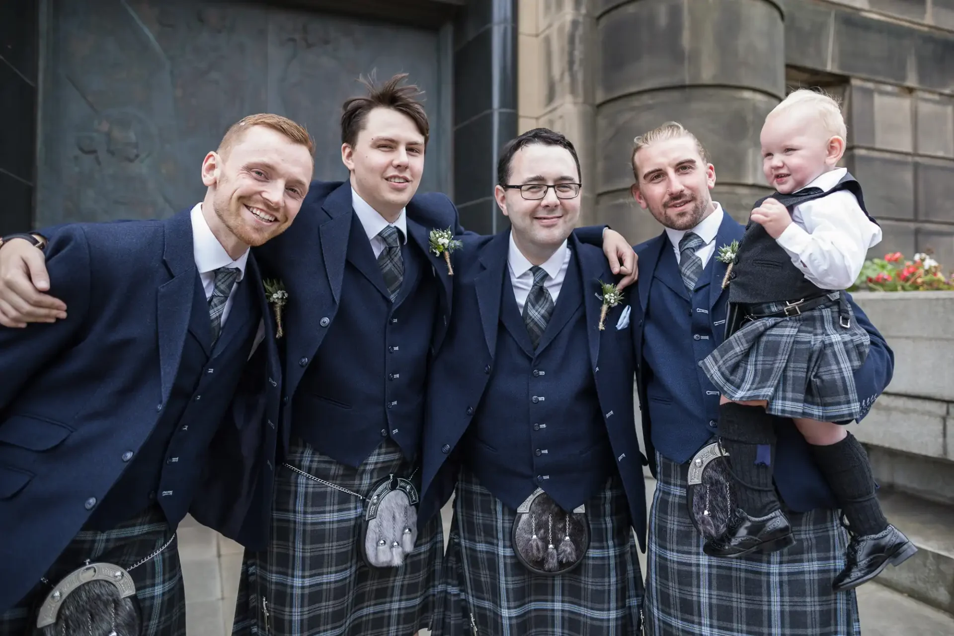 Four men and a young boy in traditional scottish kilts smiling together at a formal event.